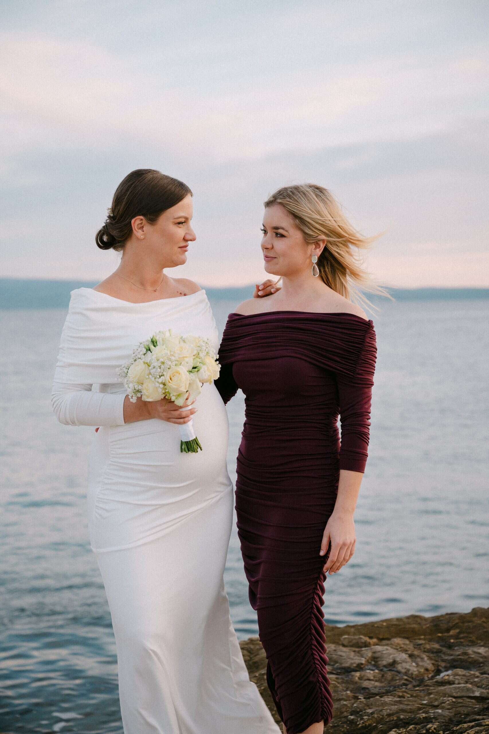 Krupni kadar mladenke i kume u elegantnim haljinama uz more, hvatajući miran trenutak tijekom zalaska sunca na plaži Bačvice u Splitu.