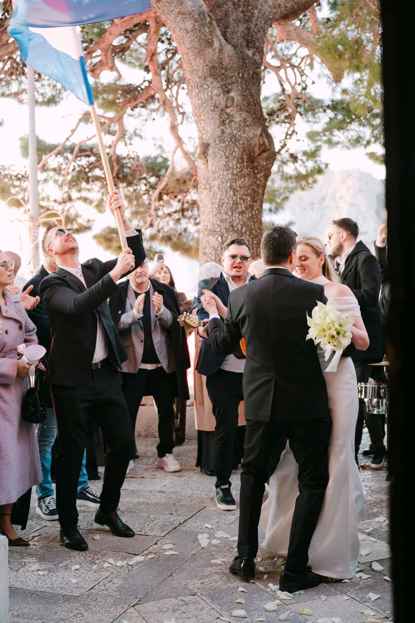 Svadbena proslava ispred crkve u Omišu gdje uzvanik drži visoko podignutu zastavu dok se mladenci vesele zajedno s gostima i plešu.