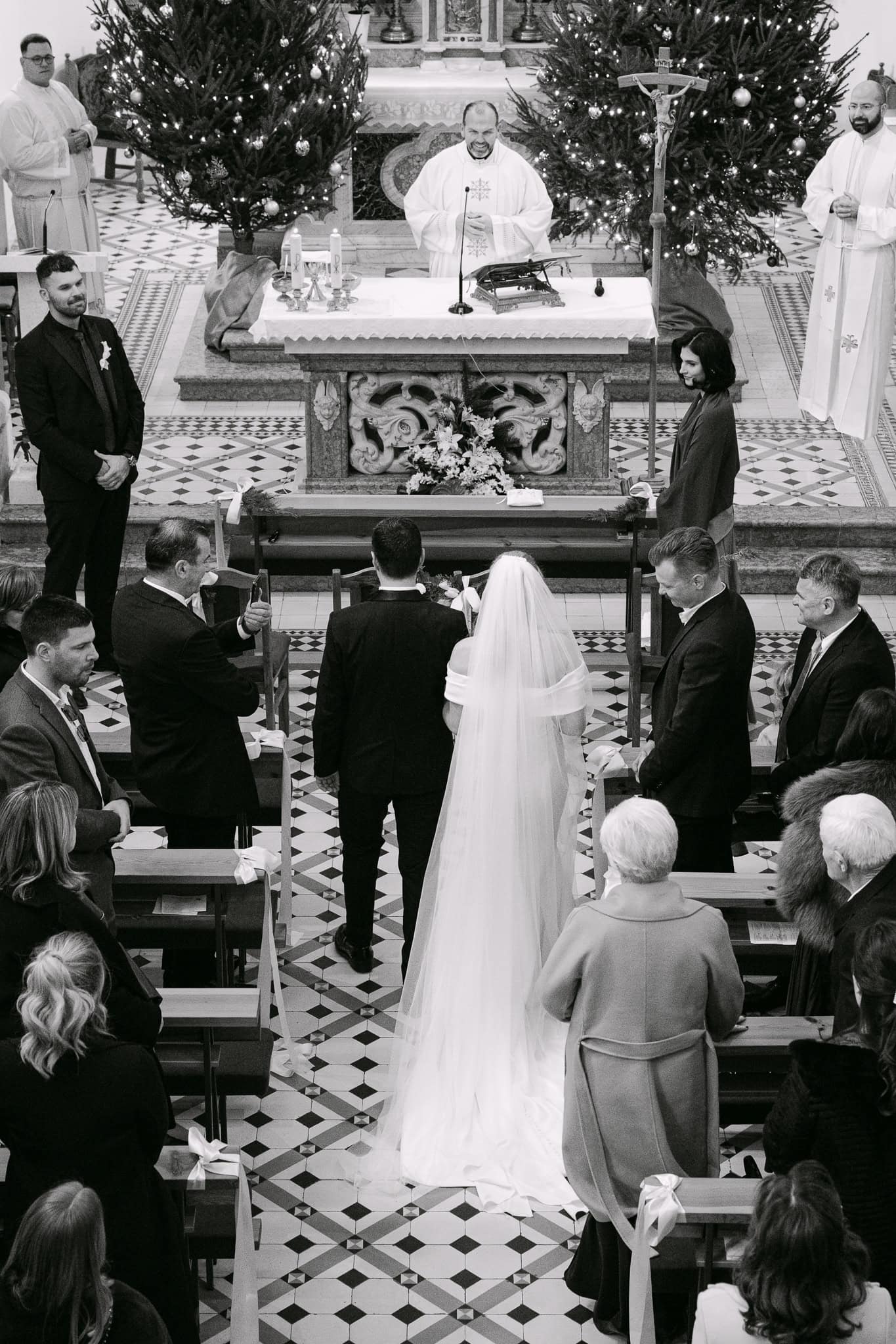 Široki crno-bijeli kadar mladenaca koji hodaju crkvenim prolazom između uzvanika prema oltaru za početak ceremonije vjenčanja.