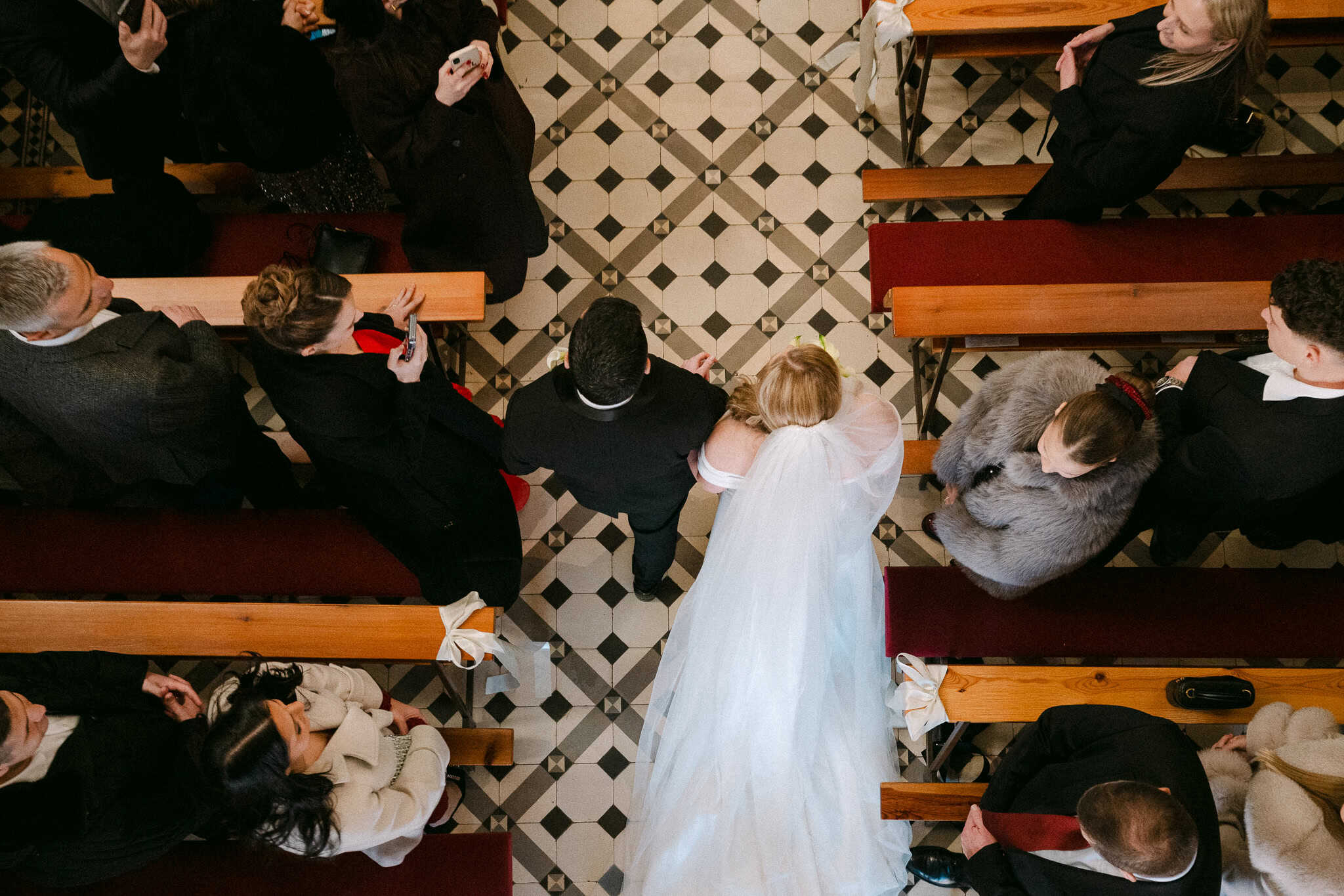Pogled odozgo na mladence koji prolaze crkvenim prolazom s crno-bijelim pločicama dok ih gosti prate pogledima iz drvenih klupa.