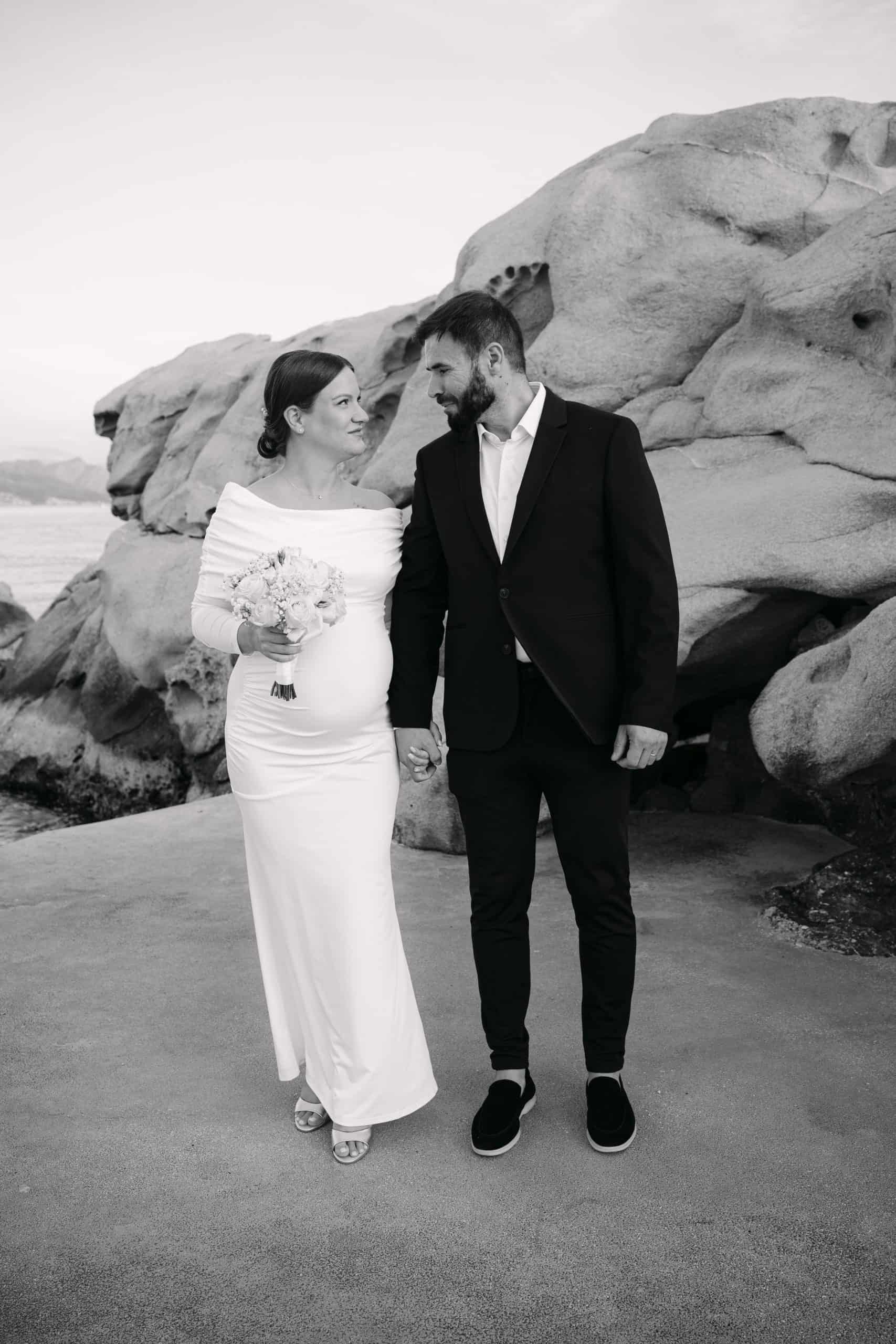 Vjenčani par na plaži s velikim kamenjem u pozadini, romantična scena.