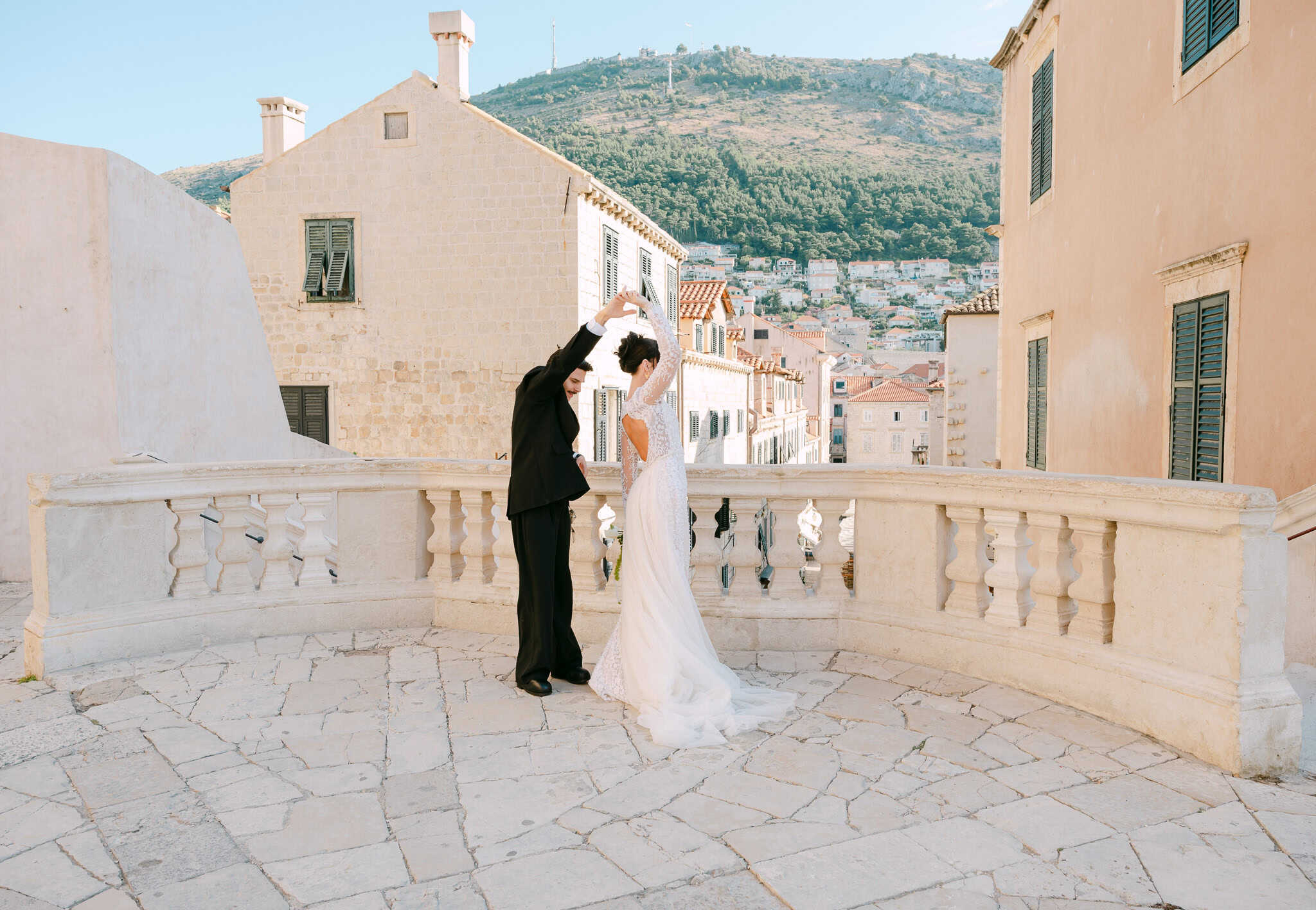 Ples mladenaca u sunčanom prijepodnevu; par se vrti na kamenoj terasi s ukrasnom balustradom ispred autentičnih dubrovačkih fasada i zelenog brda u daljini.