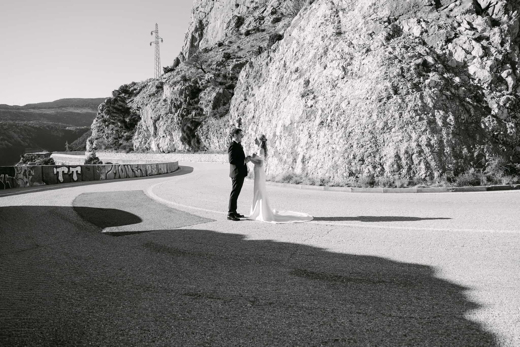 Široki crno-bijeli kadar mladenaca koji stoje na asfaltiranoj cesti ispod visoke kamene litice tijekom svadbenog sessiona u Omišu.