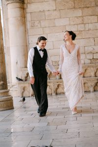 Mladenci u vjenčanici i odijelu drže se za ruke i hodaju starom kamenom ulicom tijekom svadbenog sessiona u Dubrovniku.