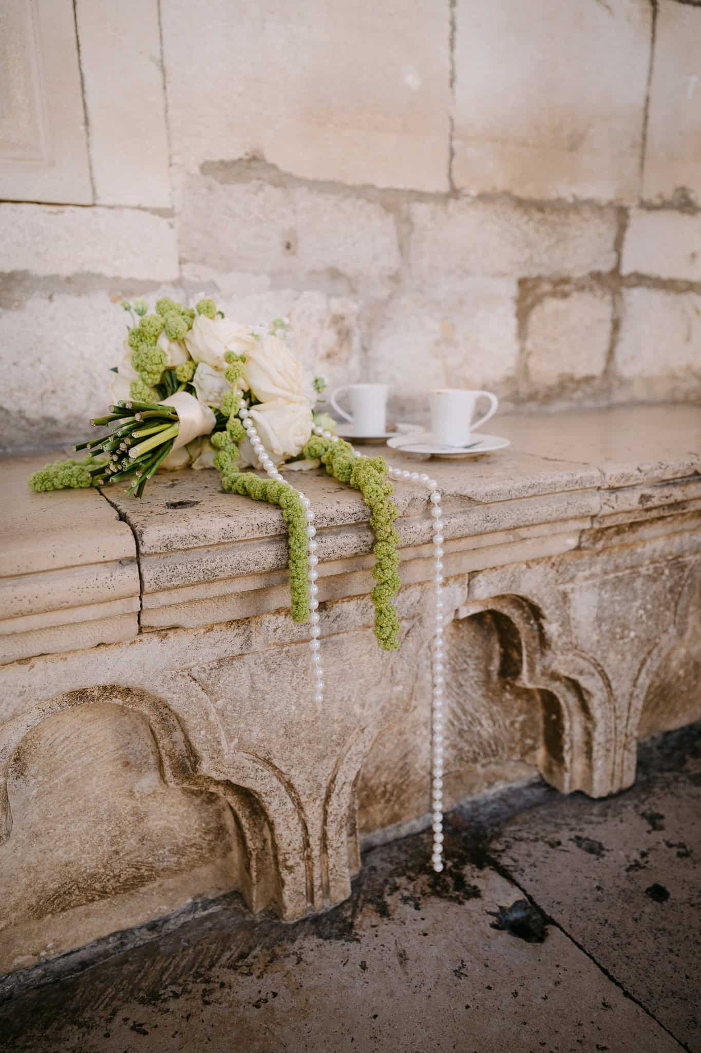 Dubrovnik, detalji vjenčanja – romantična jutarnja kava; bogati bijeli vjenčani buket s biserima položen na kamenu klupu pored dvije šalice kave.