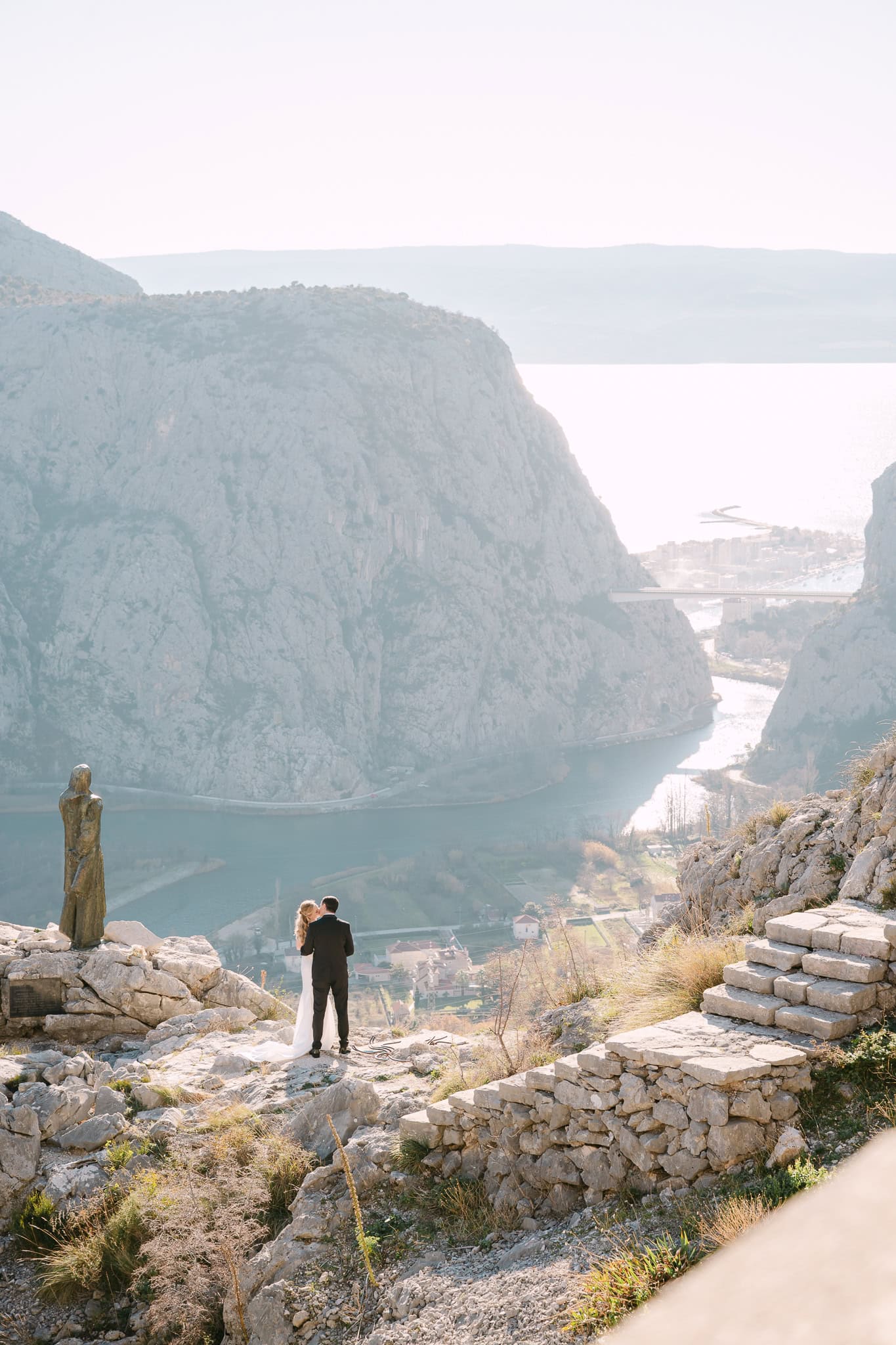 Mladenci i kip Mile Gojsalić snimljeni s leđa s panoramskim pogledom na kanjon rijeke Cetine i grad Omiš.
