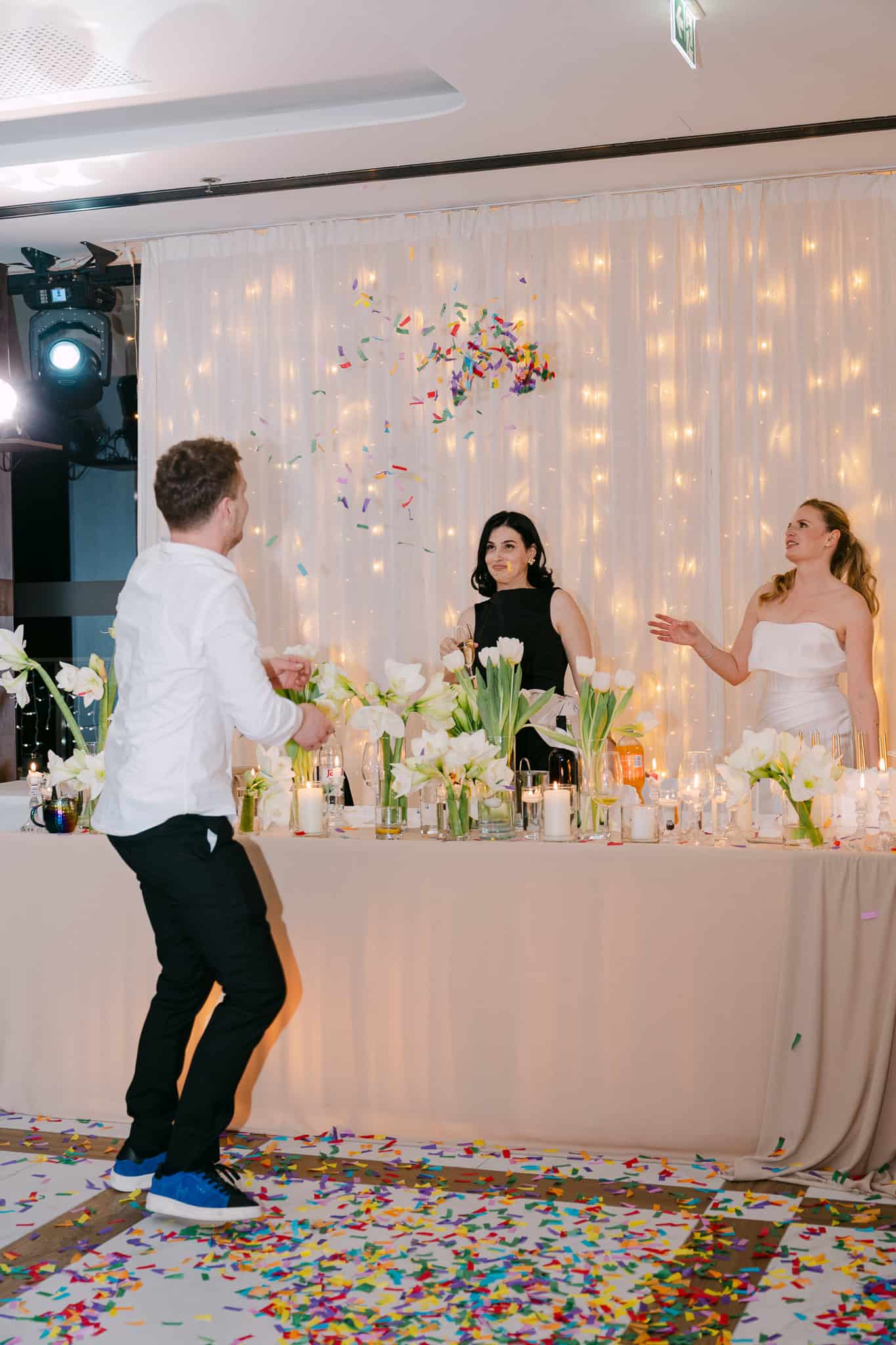 Dokumentarni kadar opuštene zabave na vjenčanju; gost veselo pleše ispred stola mladenenke i kume prepunog cvijeća dok konfeti lete zrakom, uz romantičnu pozadinu svjetlosnih lampica.