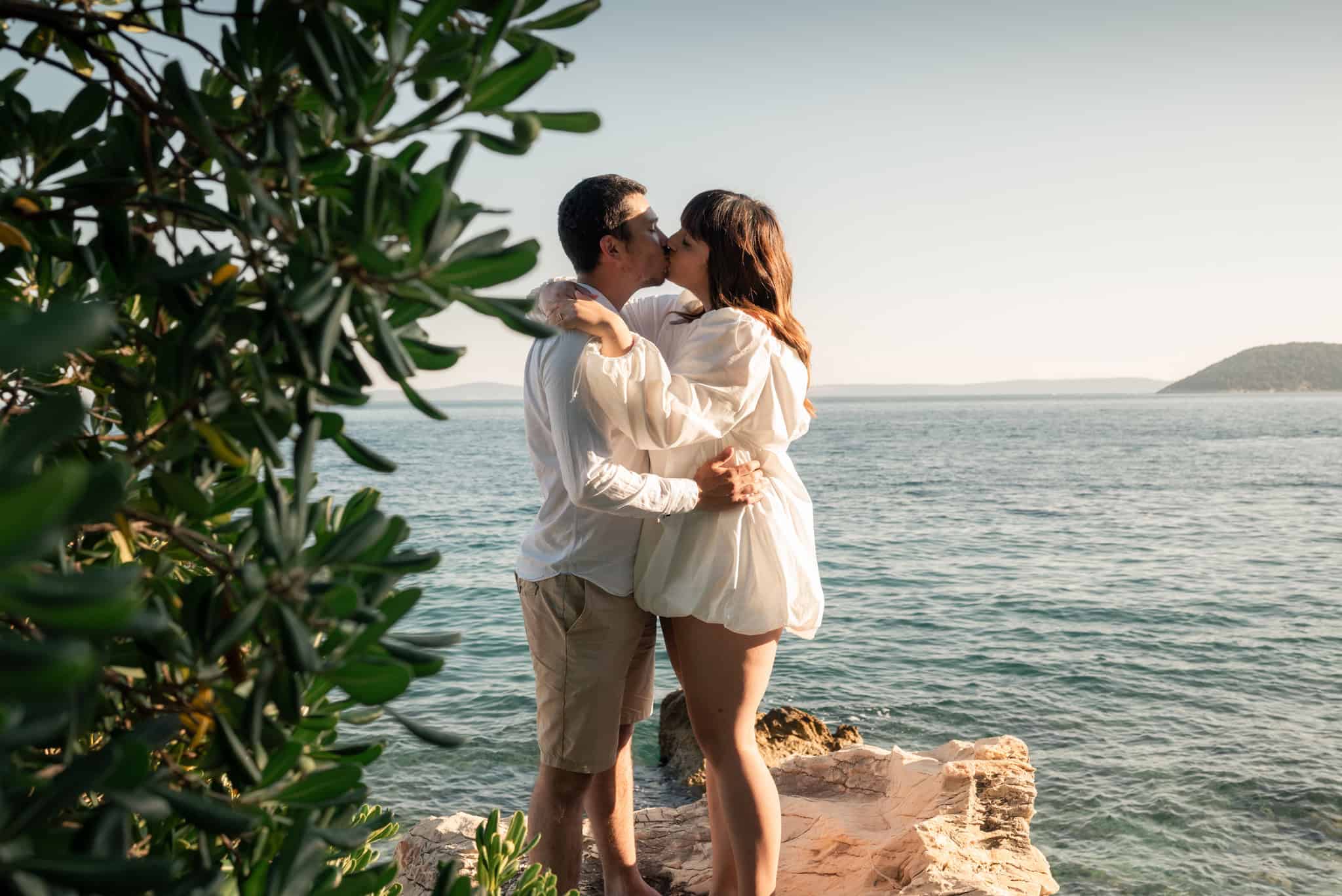 Romantični par na obali mora, ljubeći se uz zalazak sunca, okružen zelenilom i prirodom.