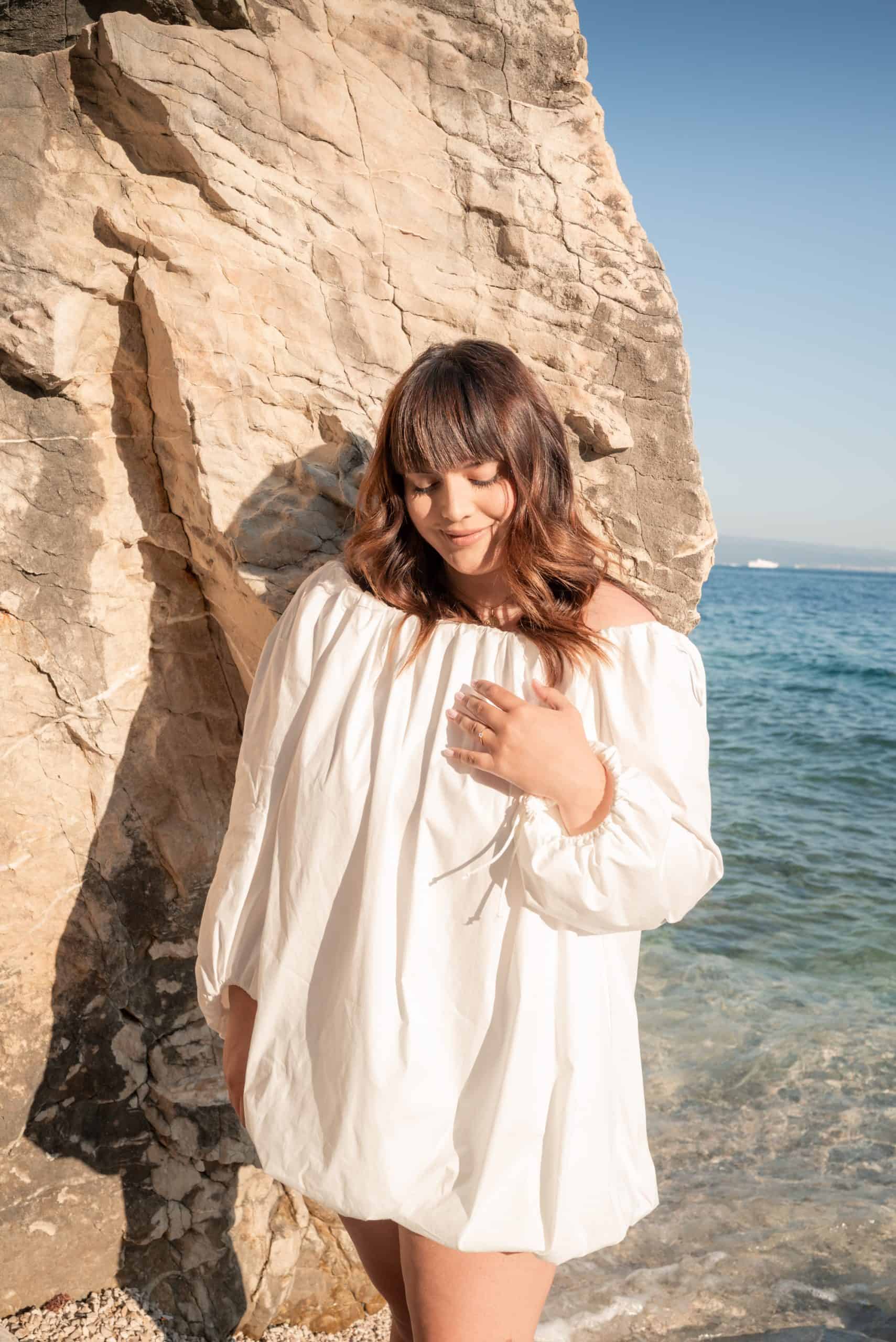 Ljetni portret buduće mladenke Petre u bijeloj haljini pored stijene na obali mora.
