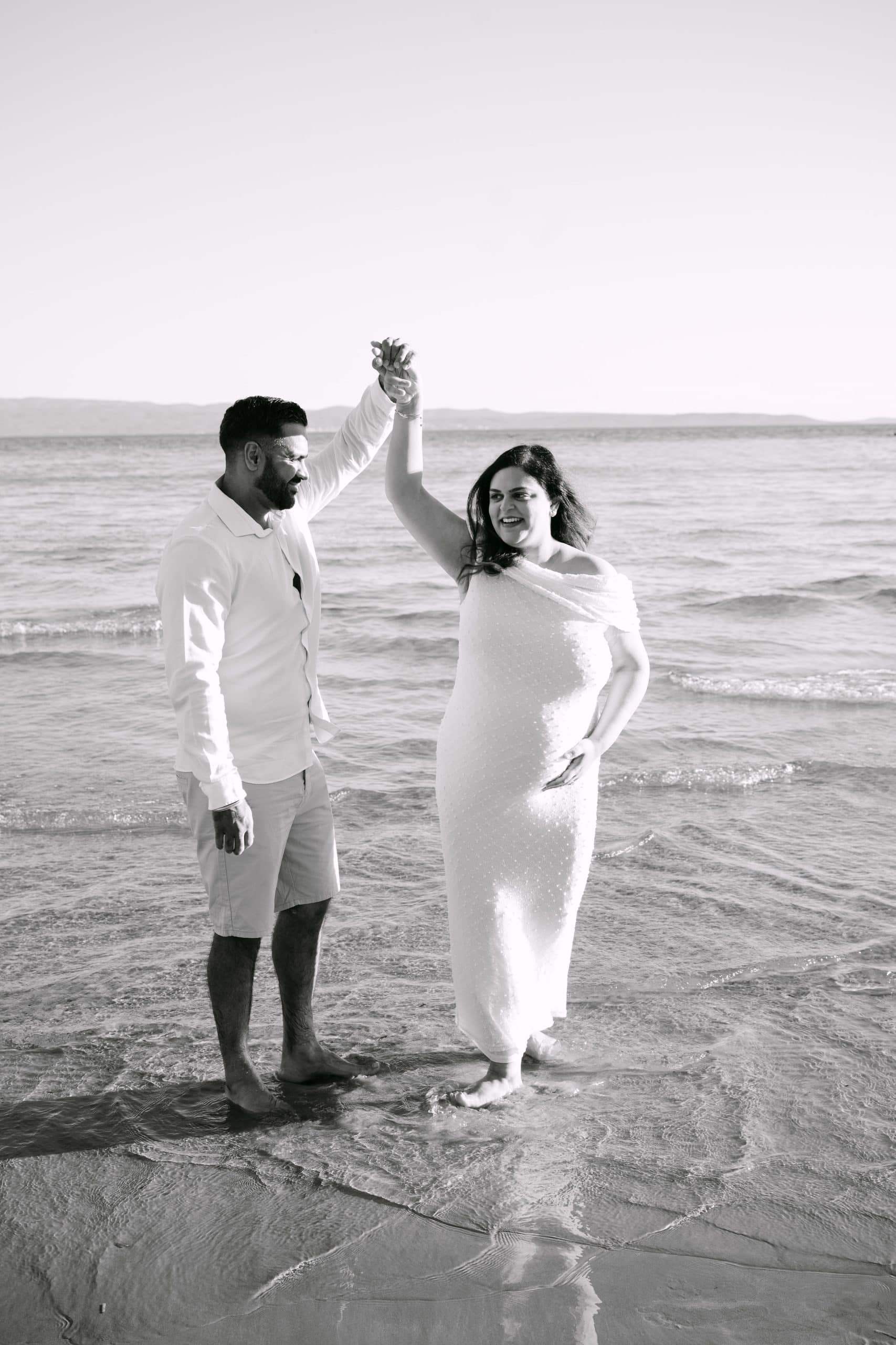 Trudnica i partner plešu na obali, slaveći trudnoću na plaži.