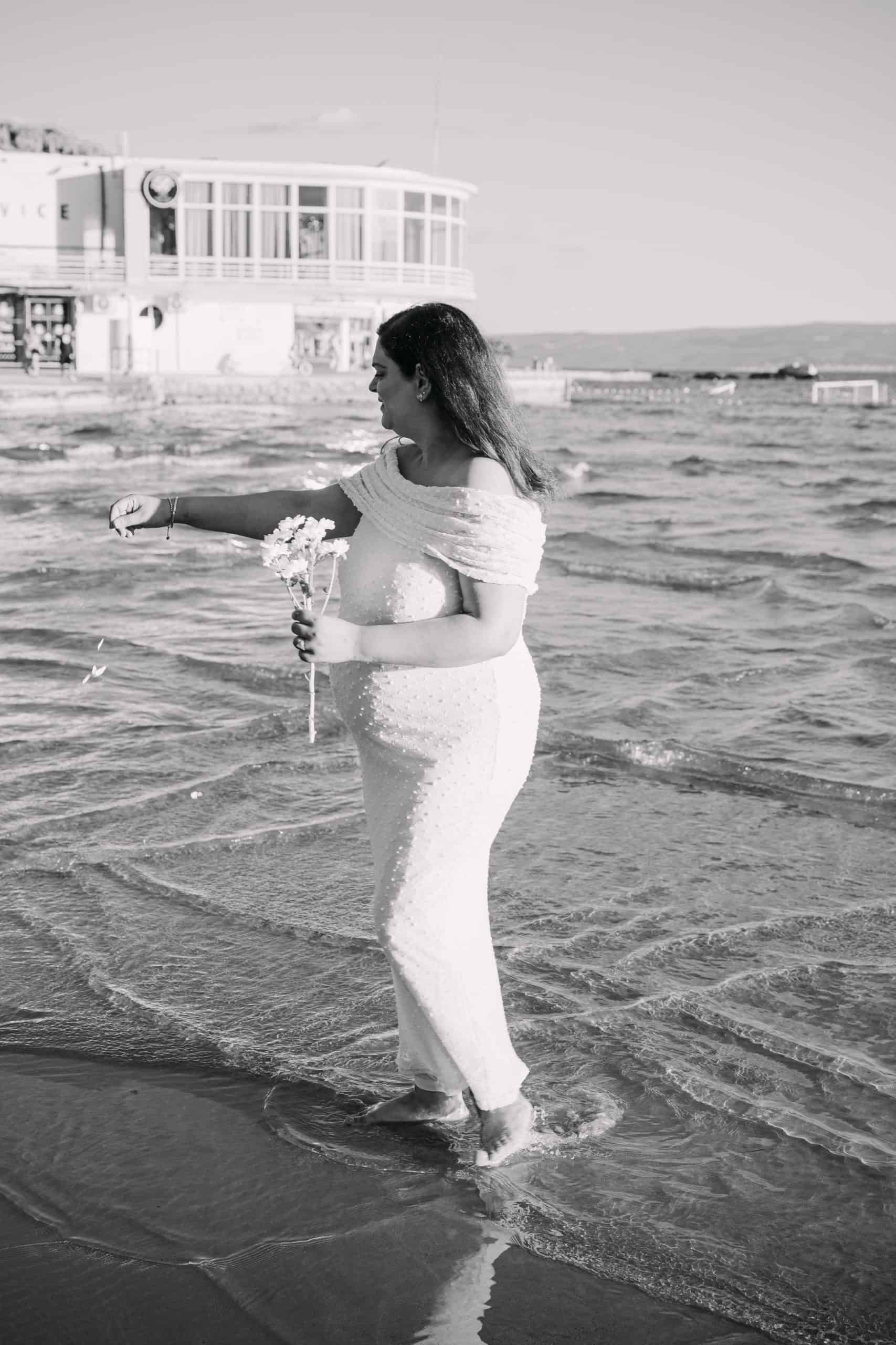 Trudna žena u bijeloj haljini stoji u plitkoj vodi na plaži, držeći cvijeće.