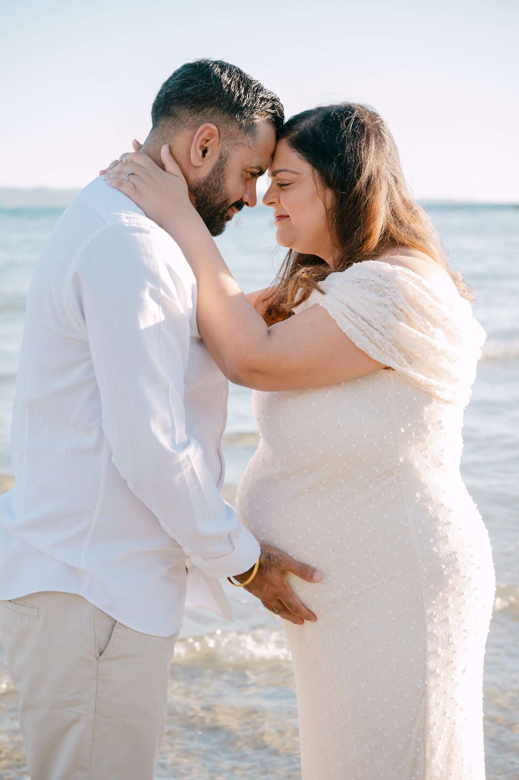 Trudnica i partner se grle na plaži, slaveći trudnoću i ljubav.