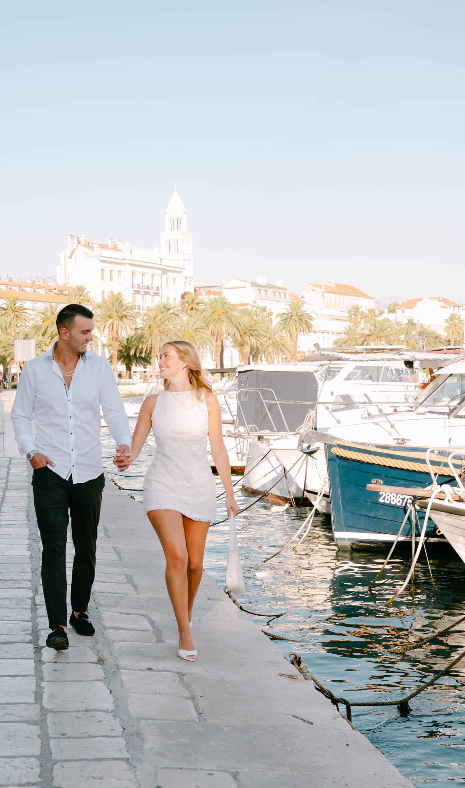 Zaručeni par šeta rivom s brodovima i povijesnom arhitekturom u pozadini.