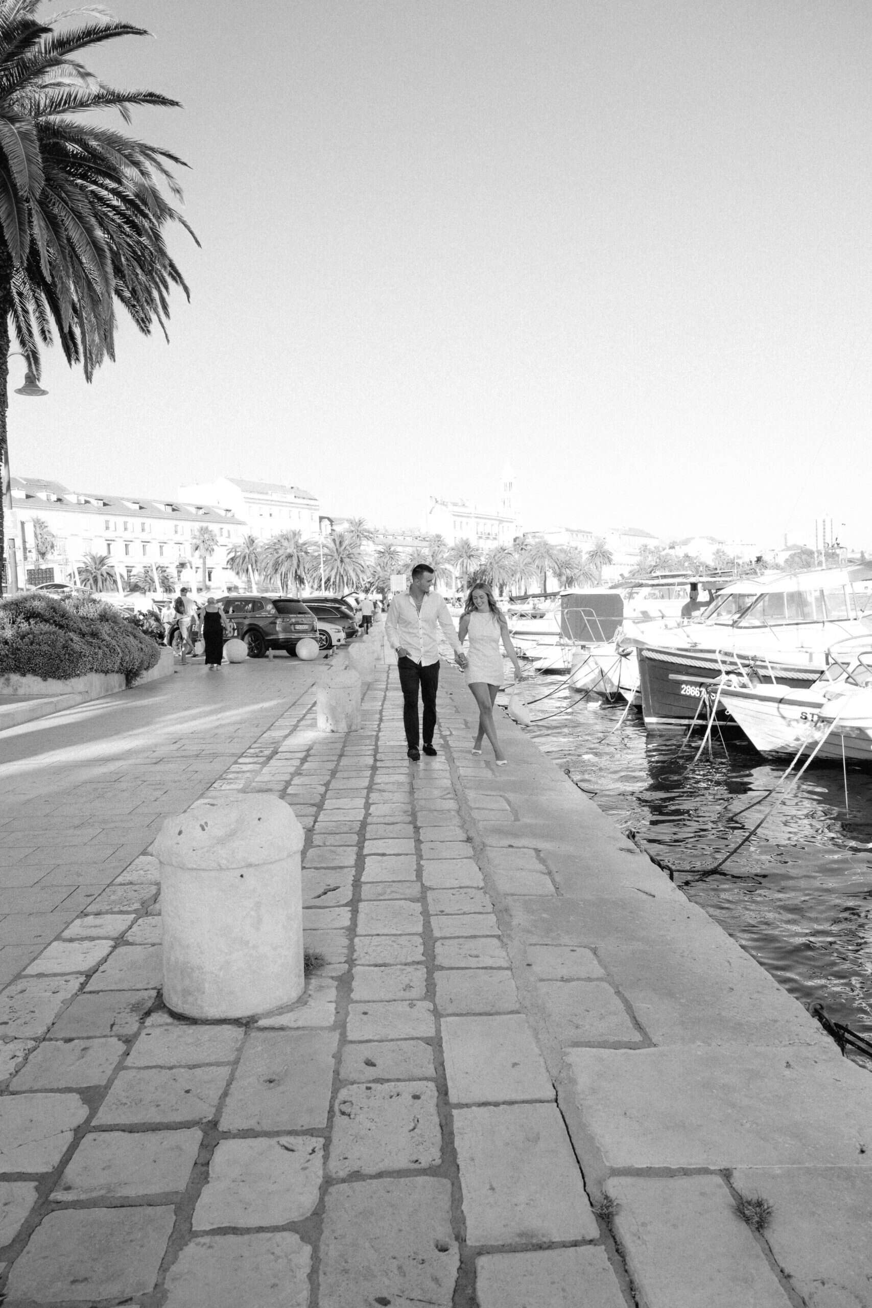Par šeta uz obalu s brodovima i palmama u pozadini, crno-bijela fotografija.