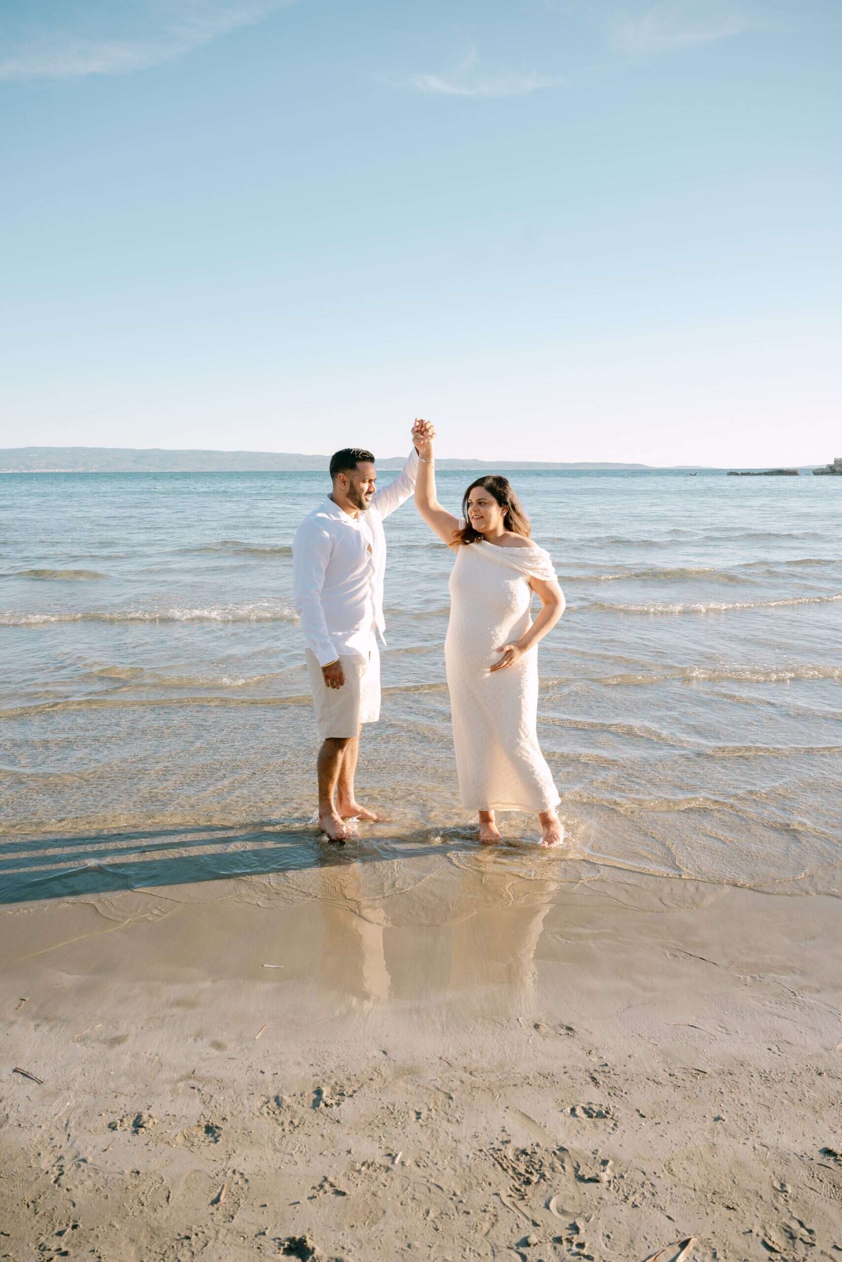 Trudna žena i partner se drži za ruke i pleše na plaži.