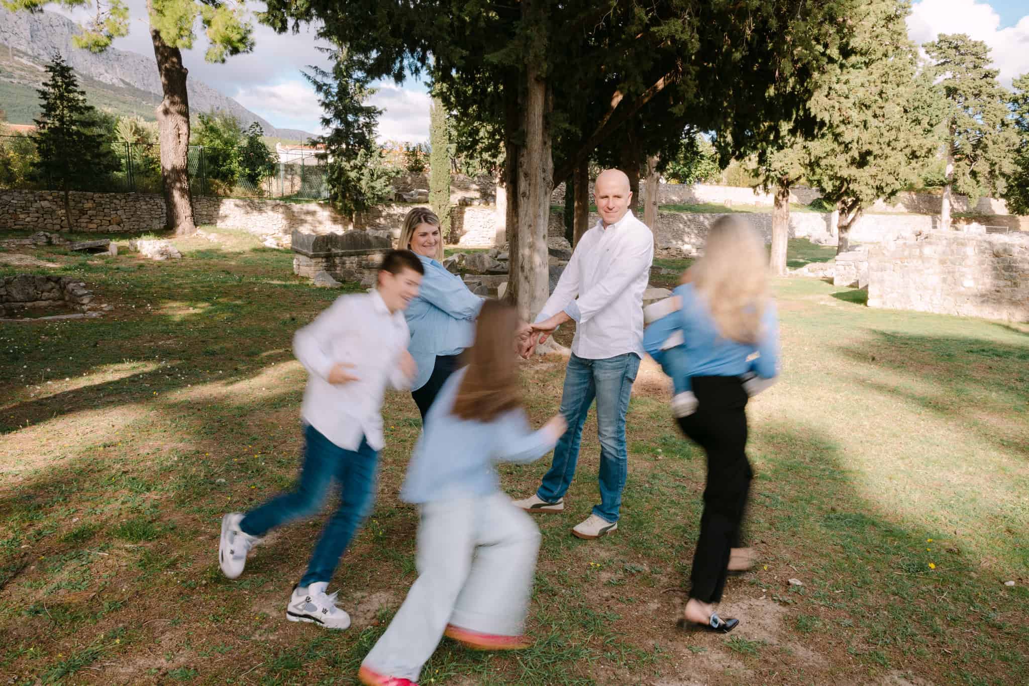 Obitelj se igra zajedno na otvorenom u parku s drvećem i povijesnim ruševinama u pozadini.