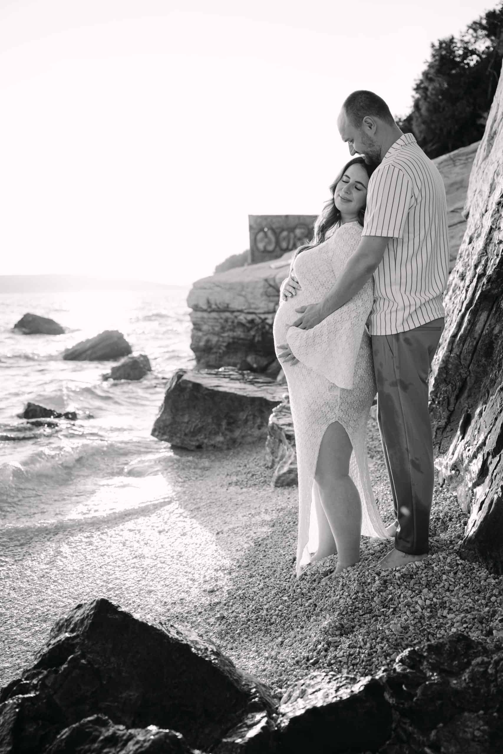 Ljubavni trudni par na plaži, romantična fotografija s morem i stijenama u pozadini.