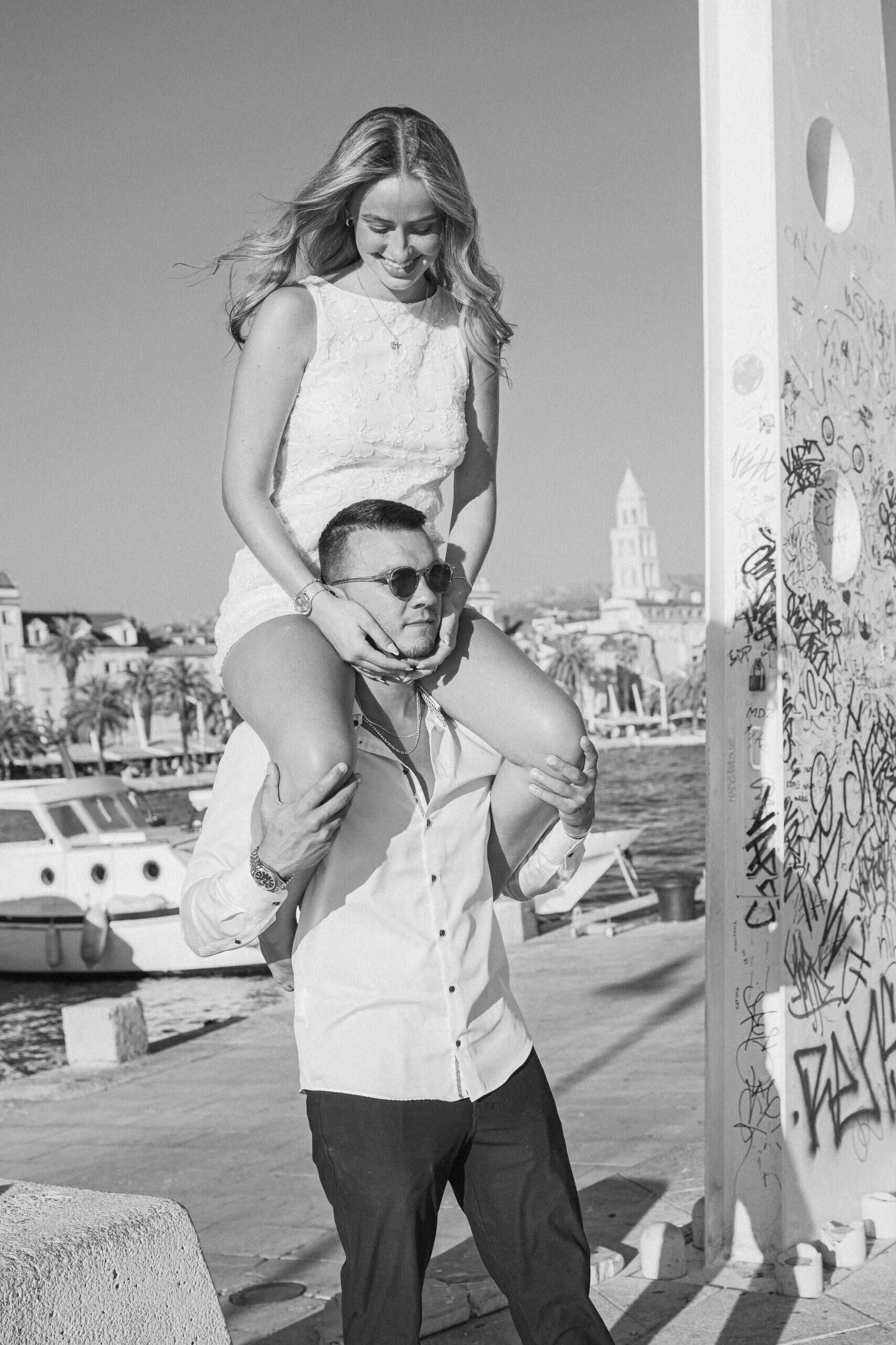 Zaručeni par u luci, žena sjedi na muškarčevim ramenima uz gradski pejzaž Splita u pozadini.