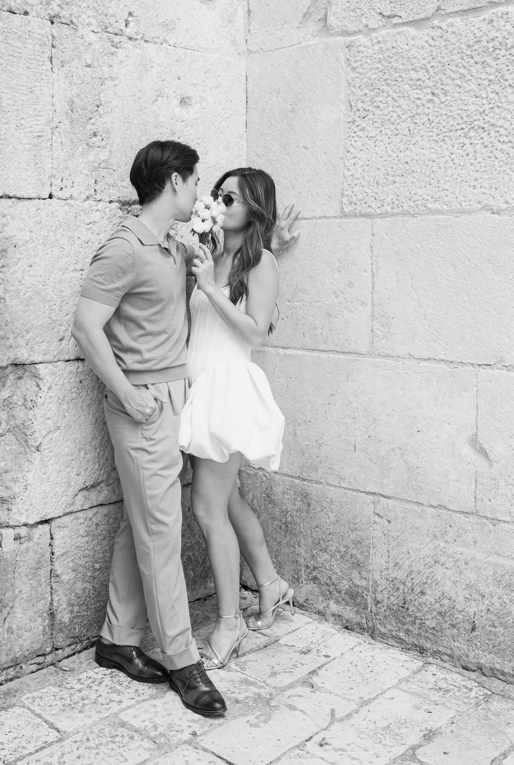 Par dijeli nježan trenutak uz kameni zid, bilježeći ljubav i intimnost u iskrenoj crno-bijeloj fotografiji.
