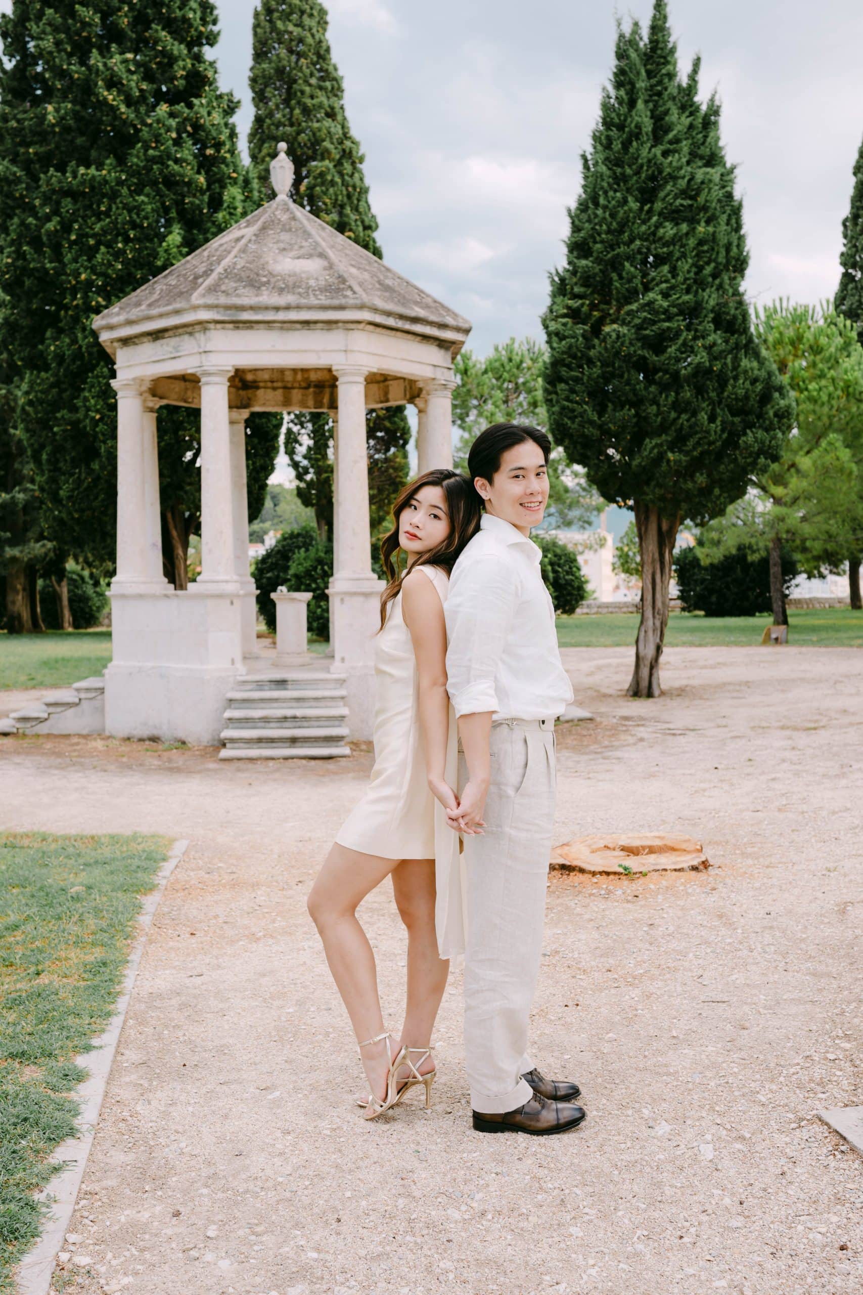 Portret vjenčanog para koji stoji leđa uz leđa u parku Sustipan u gradu Splitu s povijesnom sjenicom u pozadini.