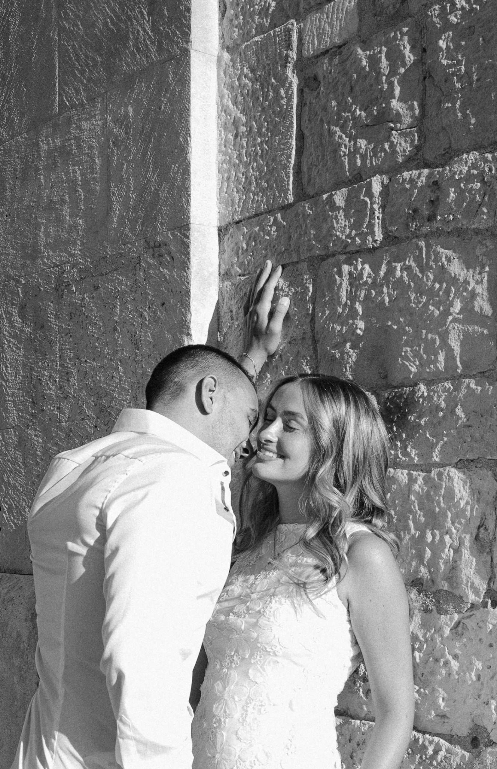 Intiman trenutak zaručenog para uz teksturirani kameni zid, crno-bijela fotografija.