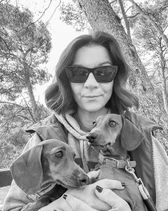 Žena s sunčanim naočalama drži dva psa u prirodi, šuma u pozadini.