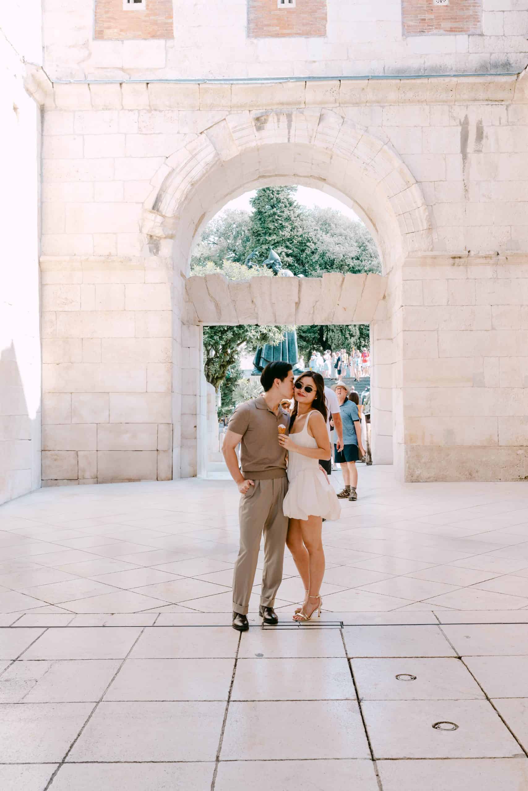 Zaljubljeni mladi par stoji ispod drevnih gradskih vrata i uživa u sunčanom danu na otvorenom.