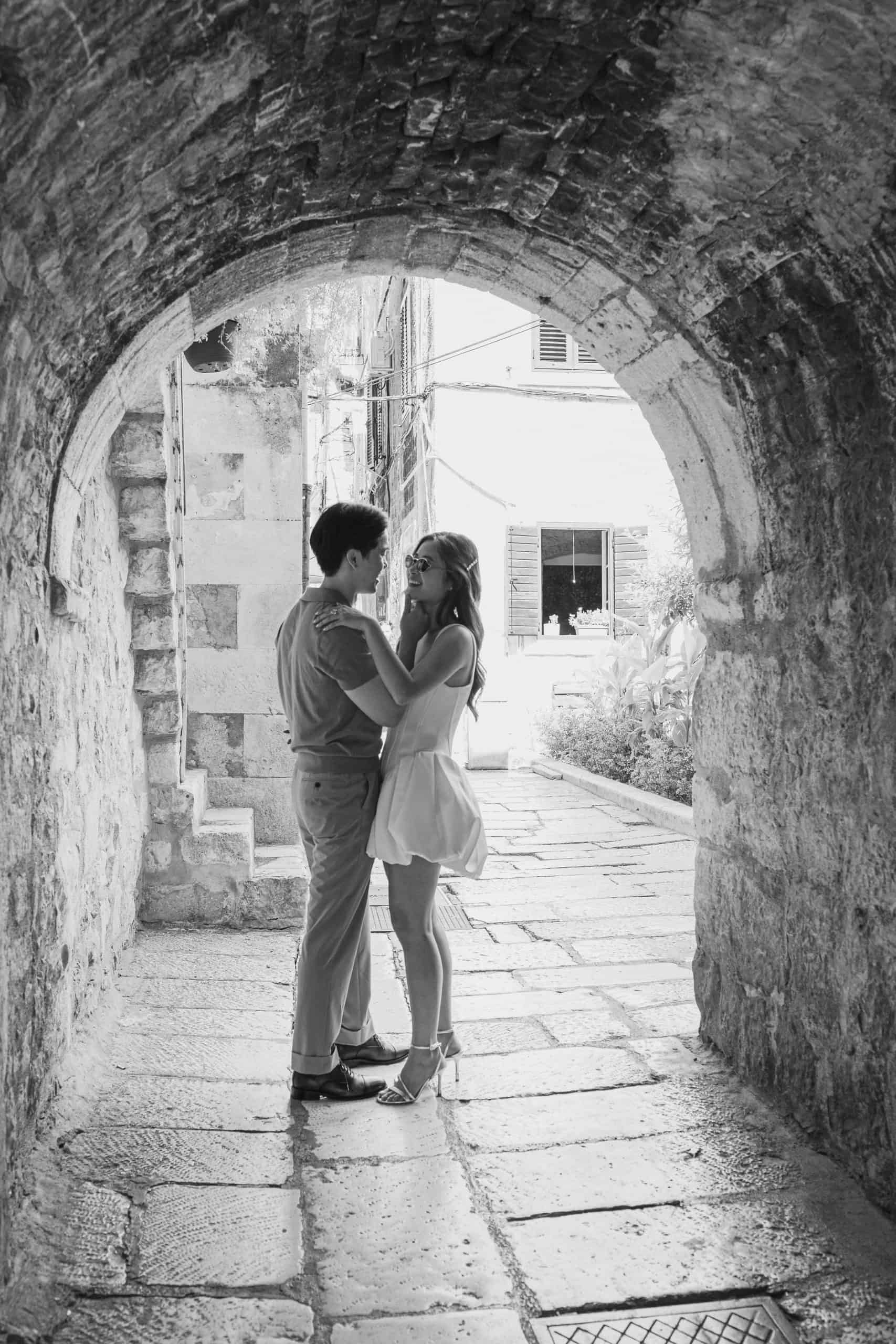 Crno-bijela fotografija para koji se grli pod lukom u povijesnom gradu.