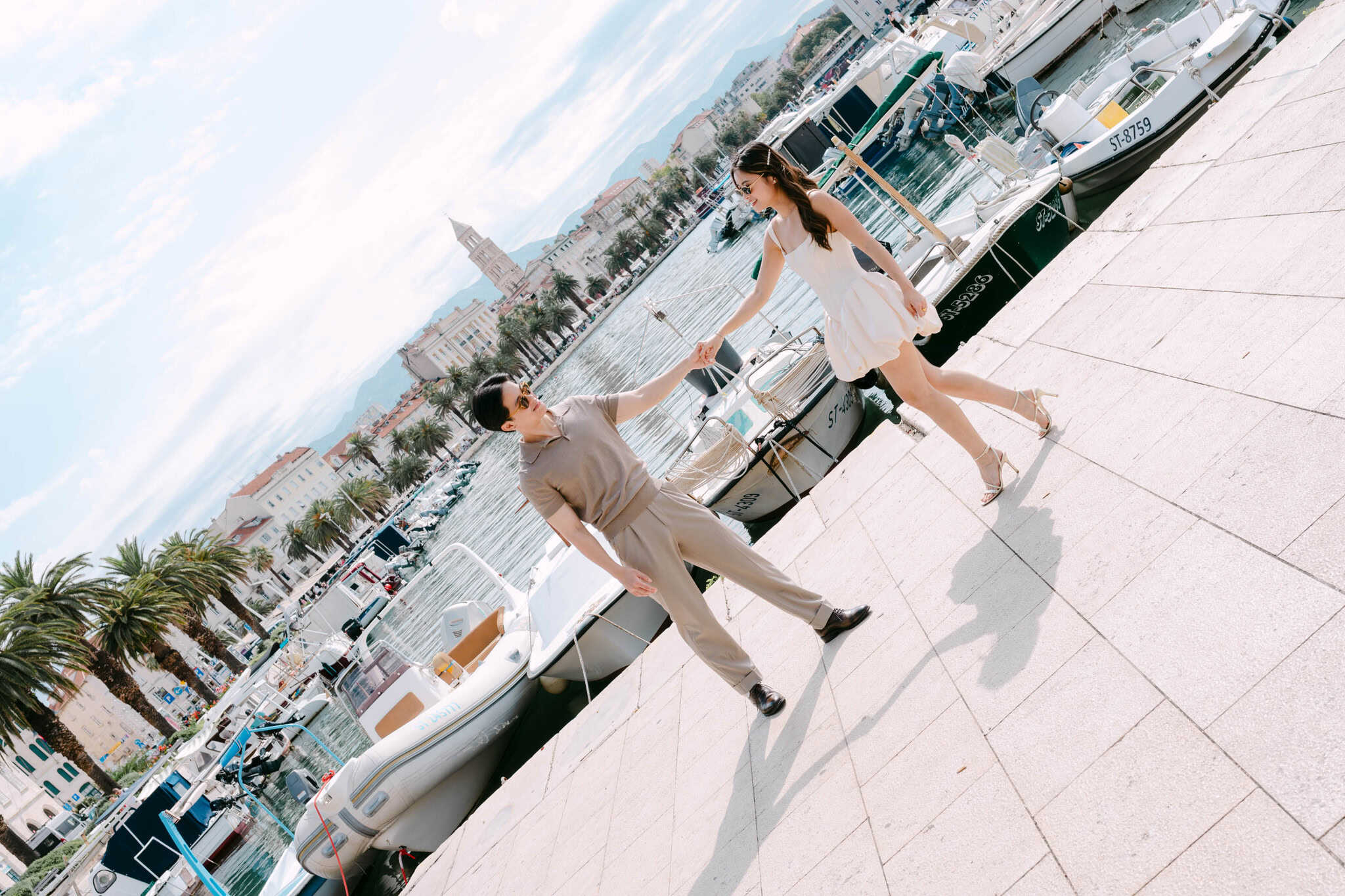 Par pleše u marini s brodovima i povijesnim gradskim pejzažom u pozadini.