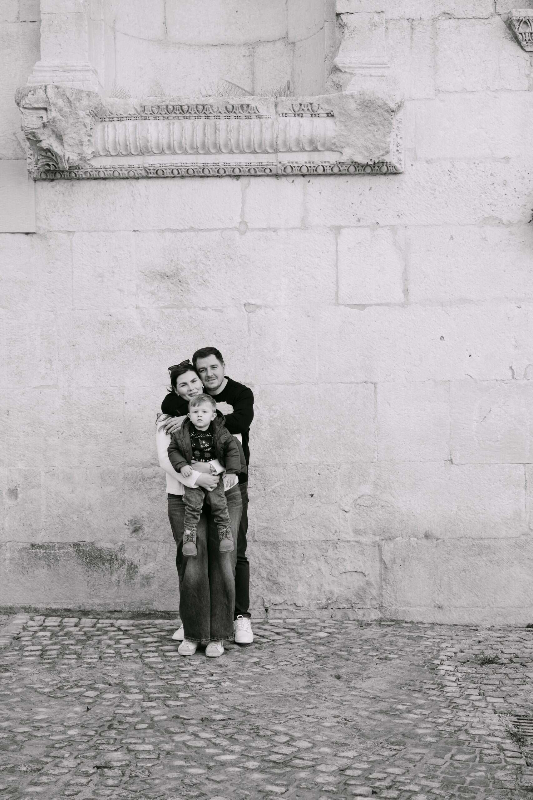 Obiteljski portret u crno-bijeloj tehnici ispred povijesne zgrade, koji bilježi iskren trenutak.