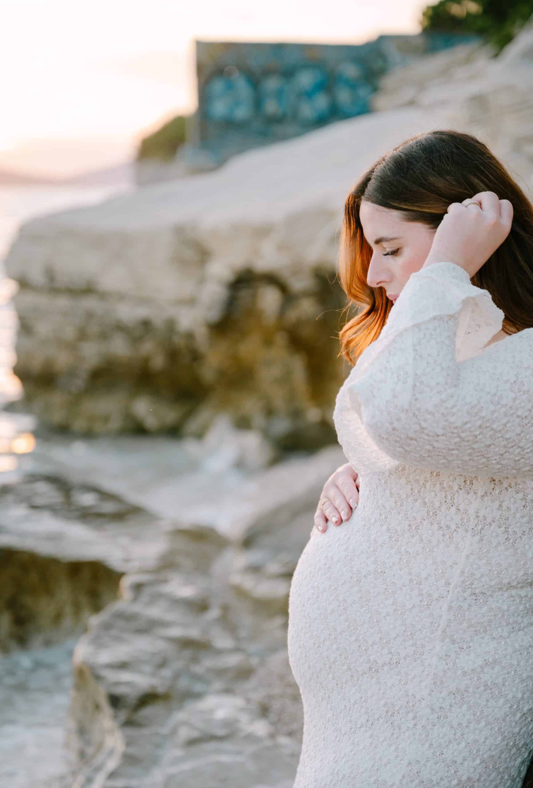 Trudnica na plaži, uživa u prirodi i miru, s pogledom na more i stijene u pozadini.