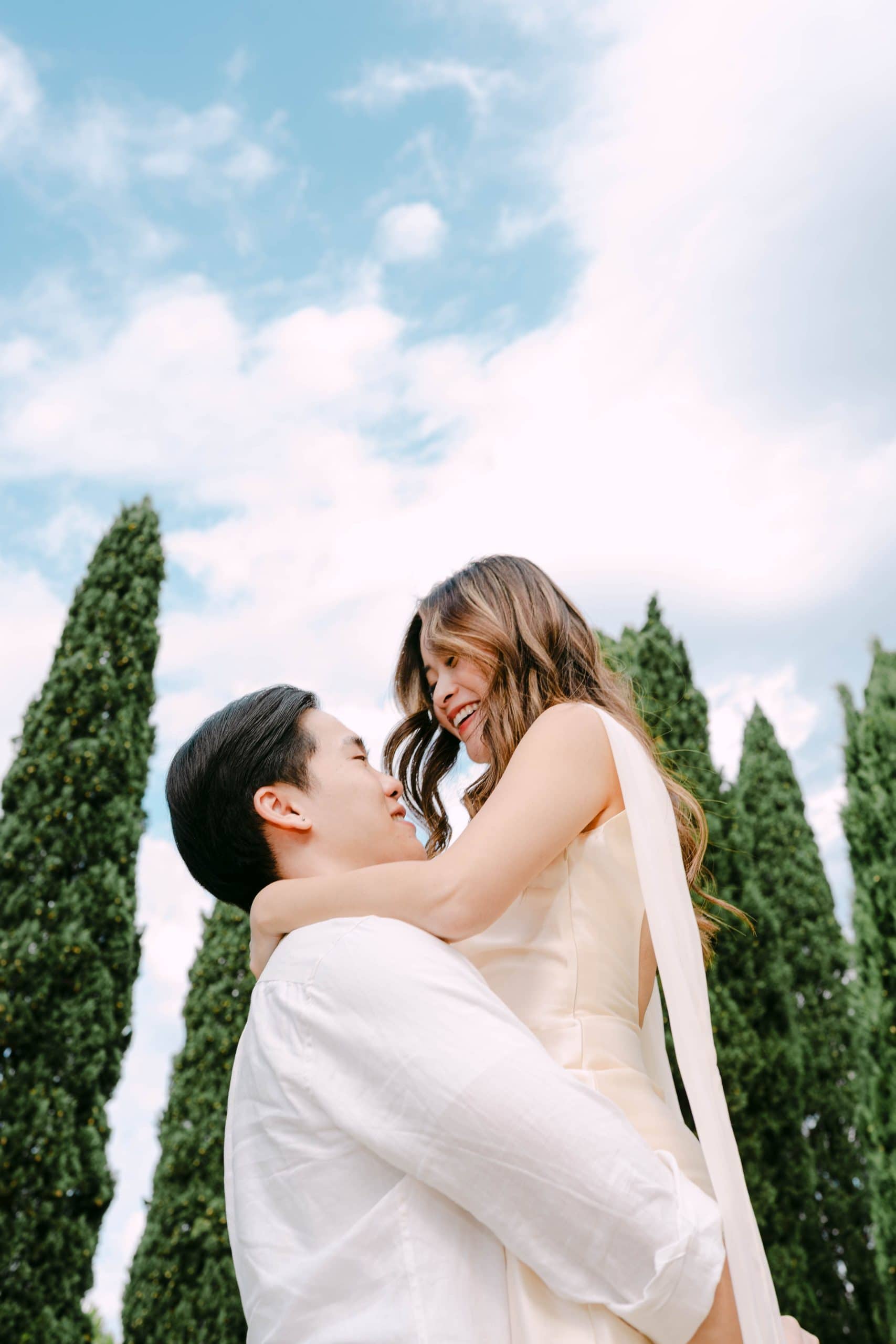 Romantični par se grli na otvorenom s visokim zelenim drvećem i vedrim nebom.