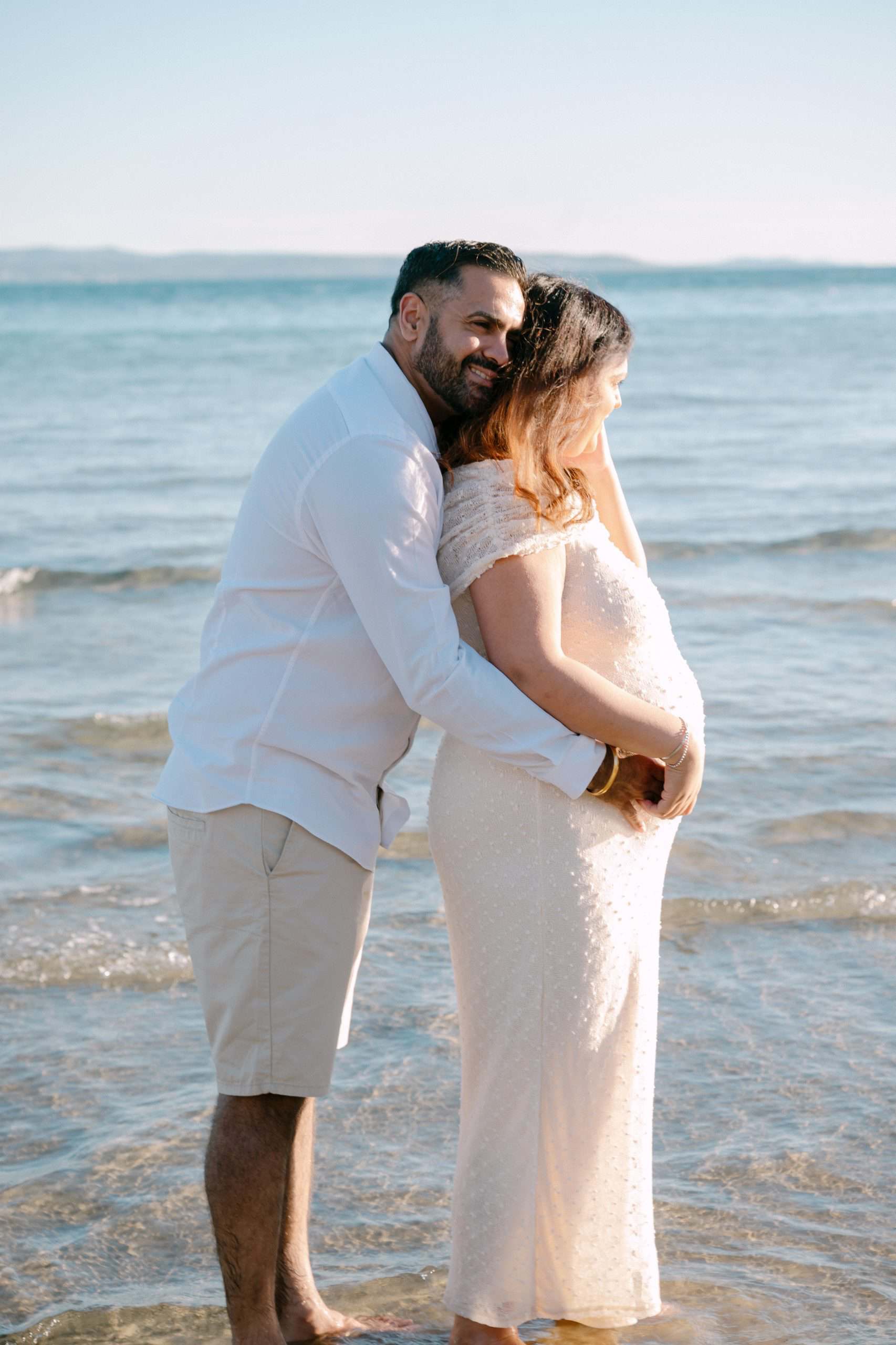 Trudnica i partner se grle na obali tijekom zalaska sunca.