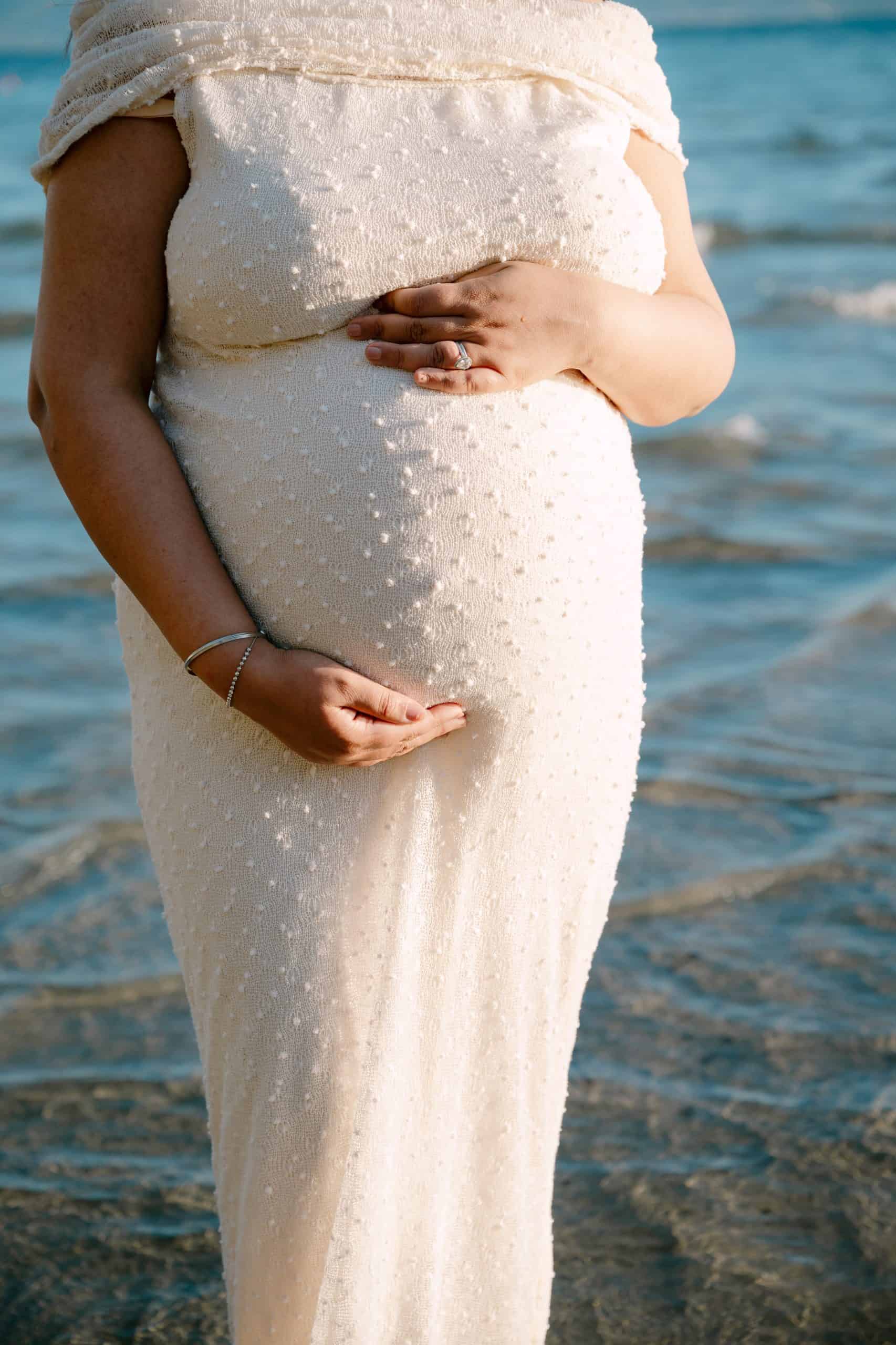 Trudnica drži svoj trbuh na plaži, uživajući u mirnom trenutku uz more.