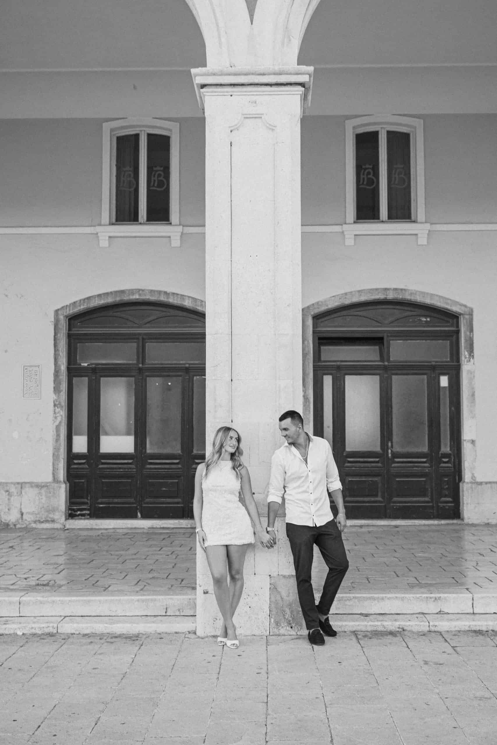 Zaručeni par se drži za ruke ispred povijesne zgrade, snimljen crno-bijelom tehnikom.