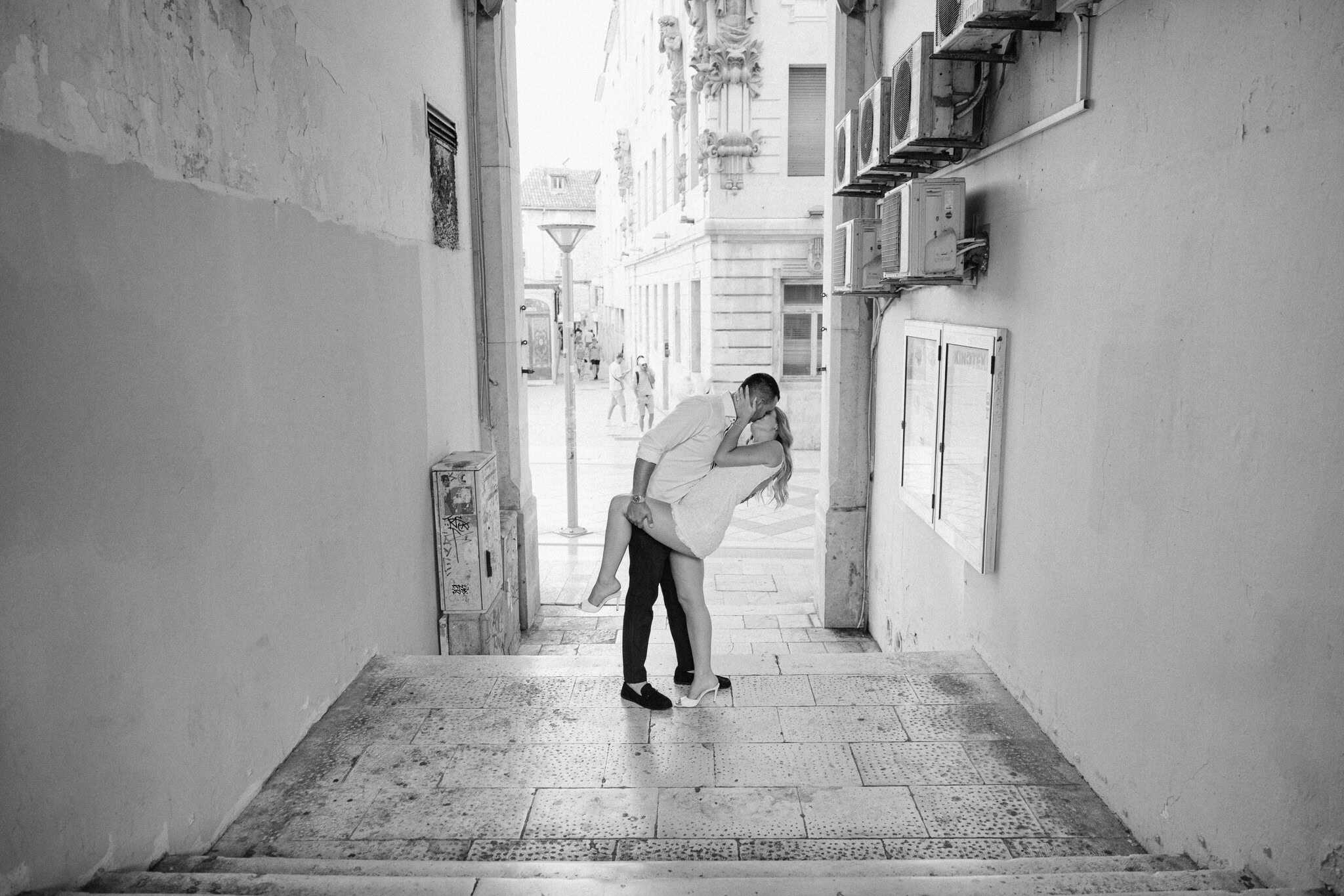 Zaručeni par se ljubi u uskoj gradskoj uličici, crno-bijela fotografija.