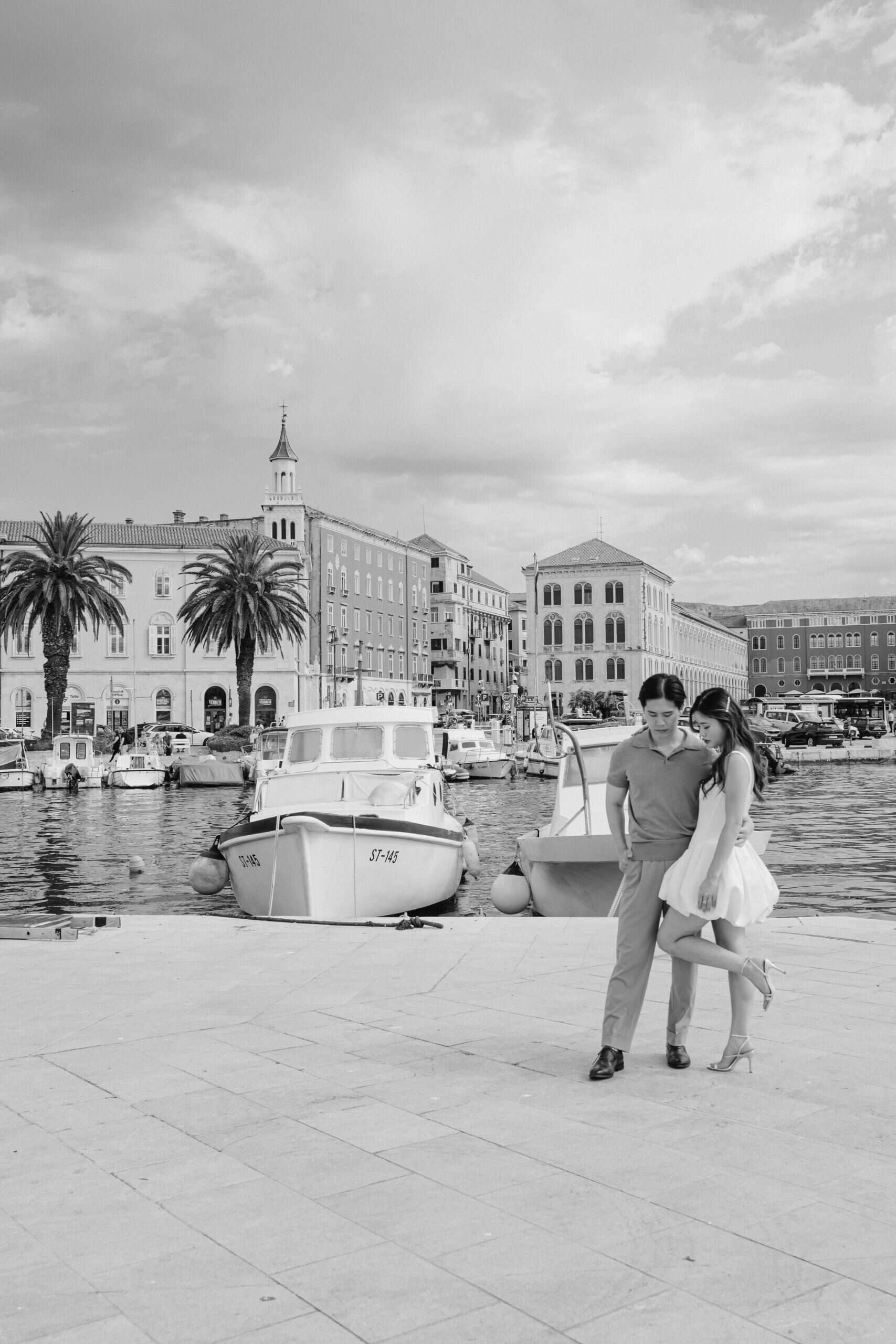 Romantični pozira pokraj marine s brodovima i povijesnim zgradama u pozadini.