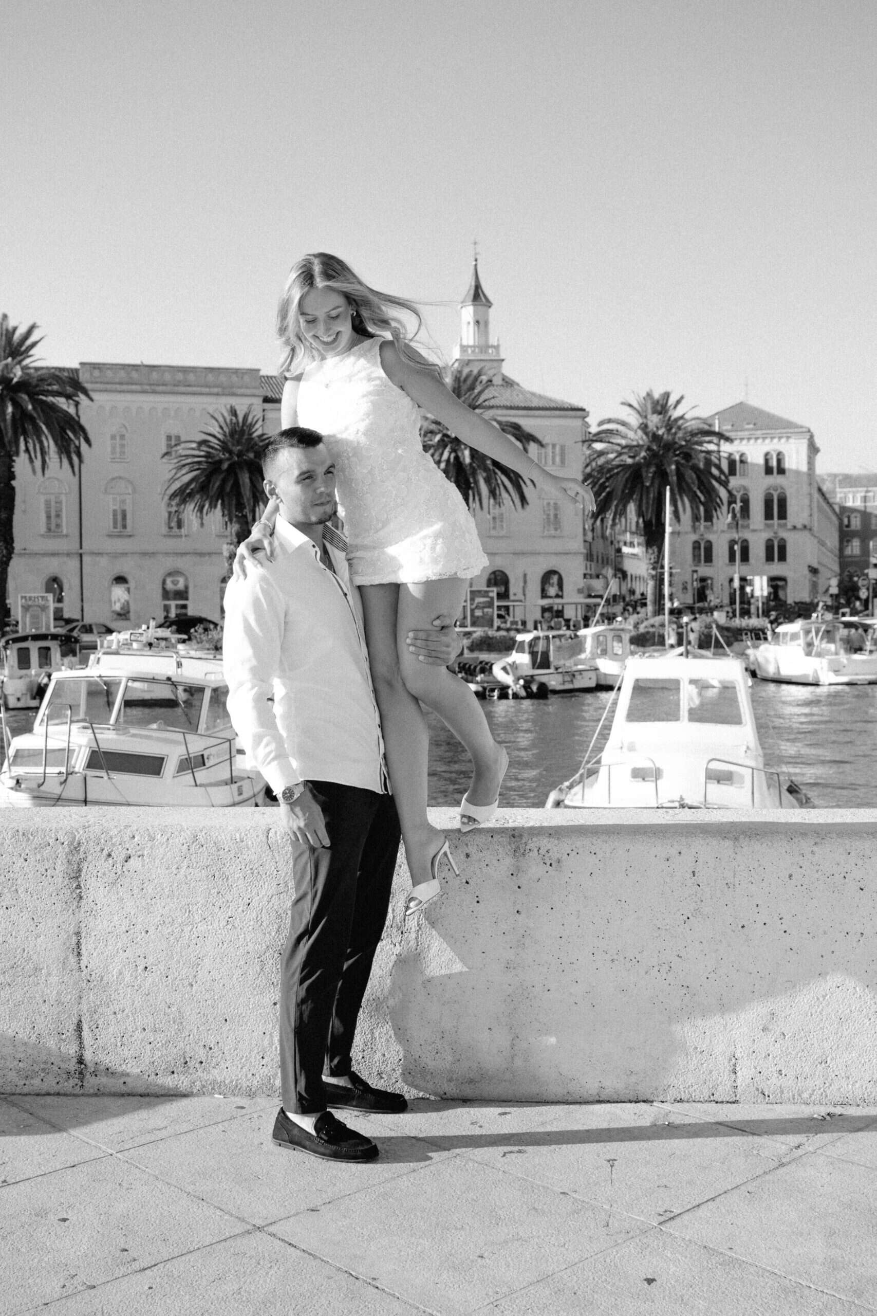 Zaručnik drži nasmijanu zaručnicu na kamenom rubu Matejuške u Splitu, crno-bijela lifestyle fotografija.