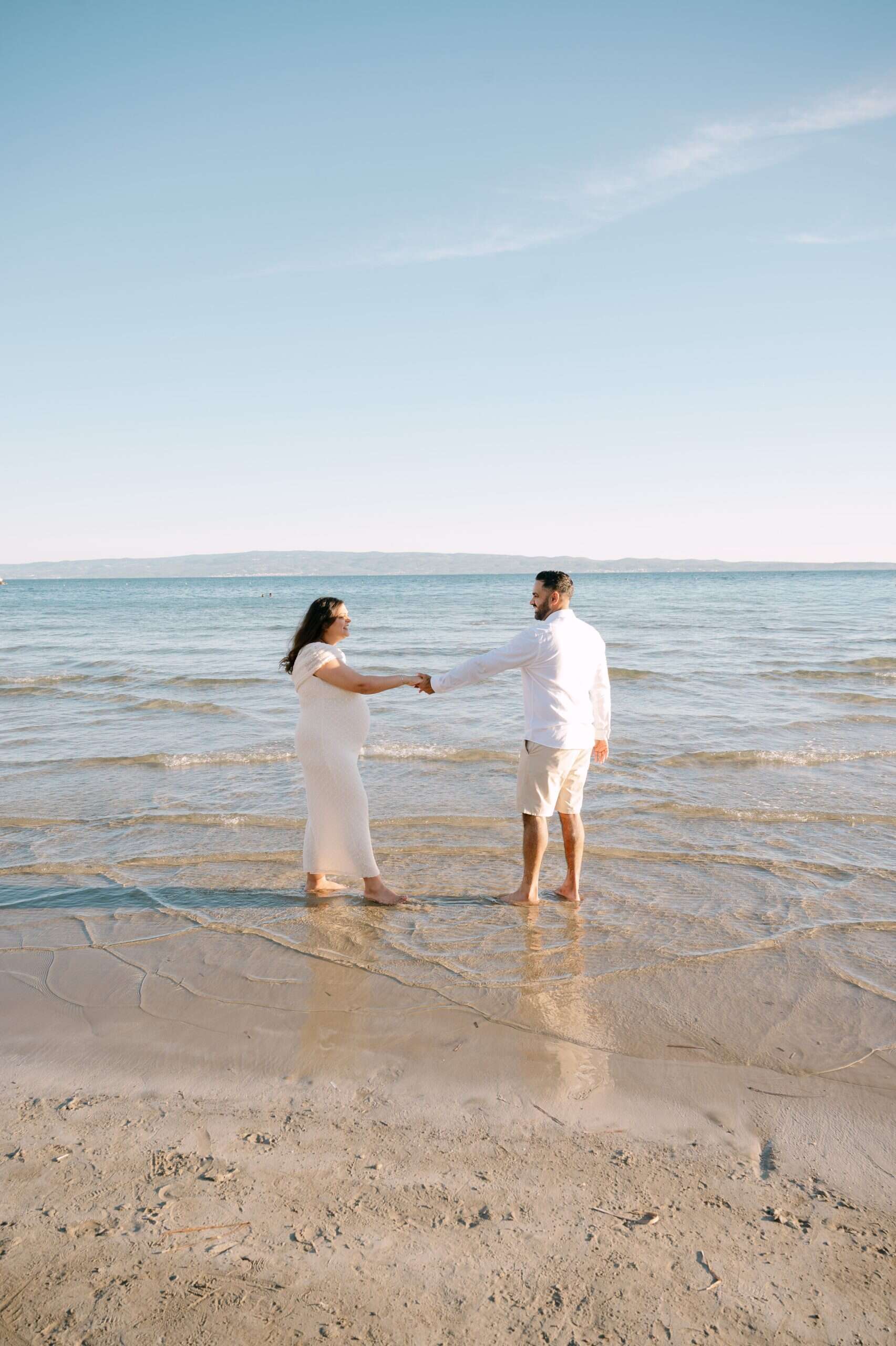 Trudnica na plaži s mužom koji se drže za ruke u plitkoj vodi.