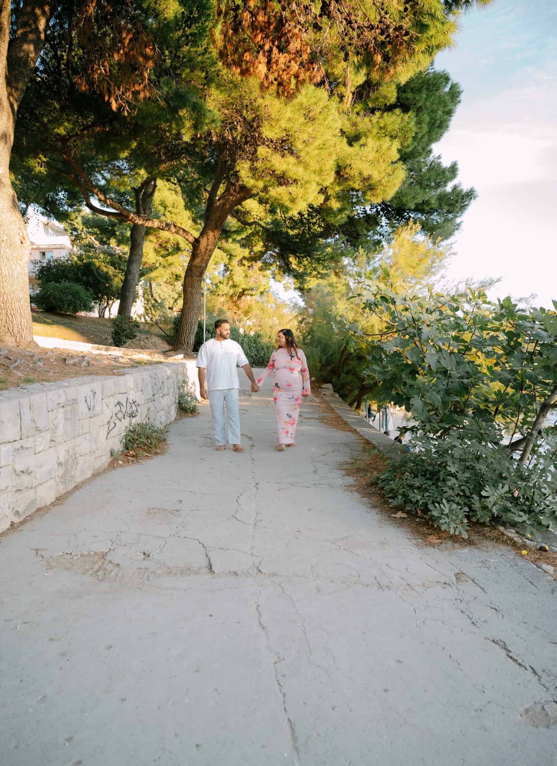 Par šeta slikovitom stazom uz more s bujnim drvećem i zelenilom.
