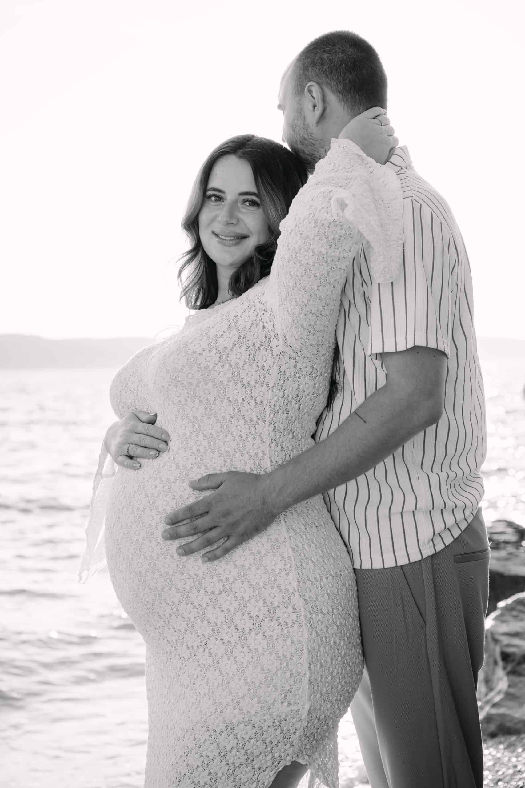 Buduća majka i suprug u zagrljaju na plaži, crno-bijela estetika ljetnog sessiona.