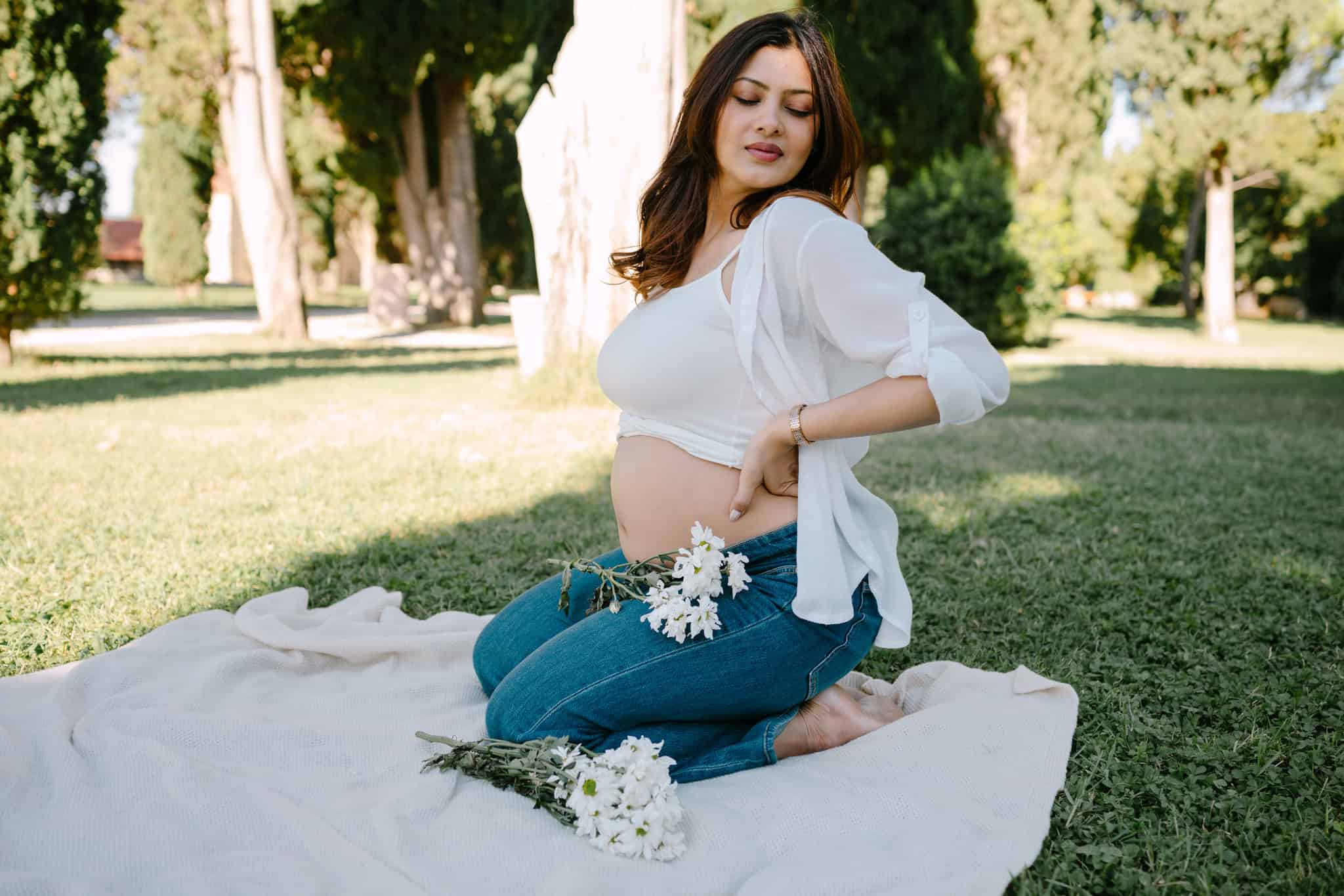 Buduća majka u bijeloj majici i plavim trapericama na travnjaku, okružena cvijećem i drvećem.