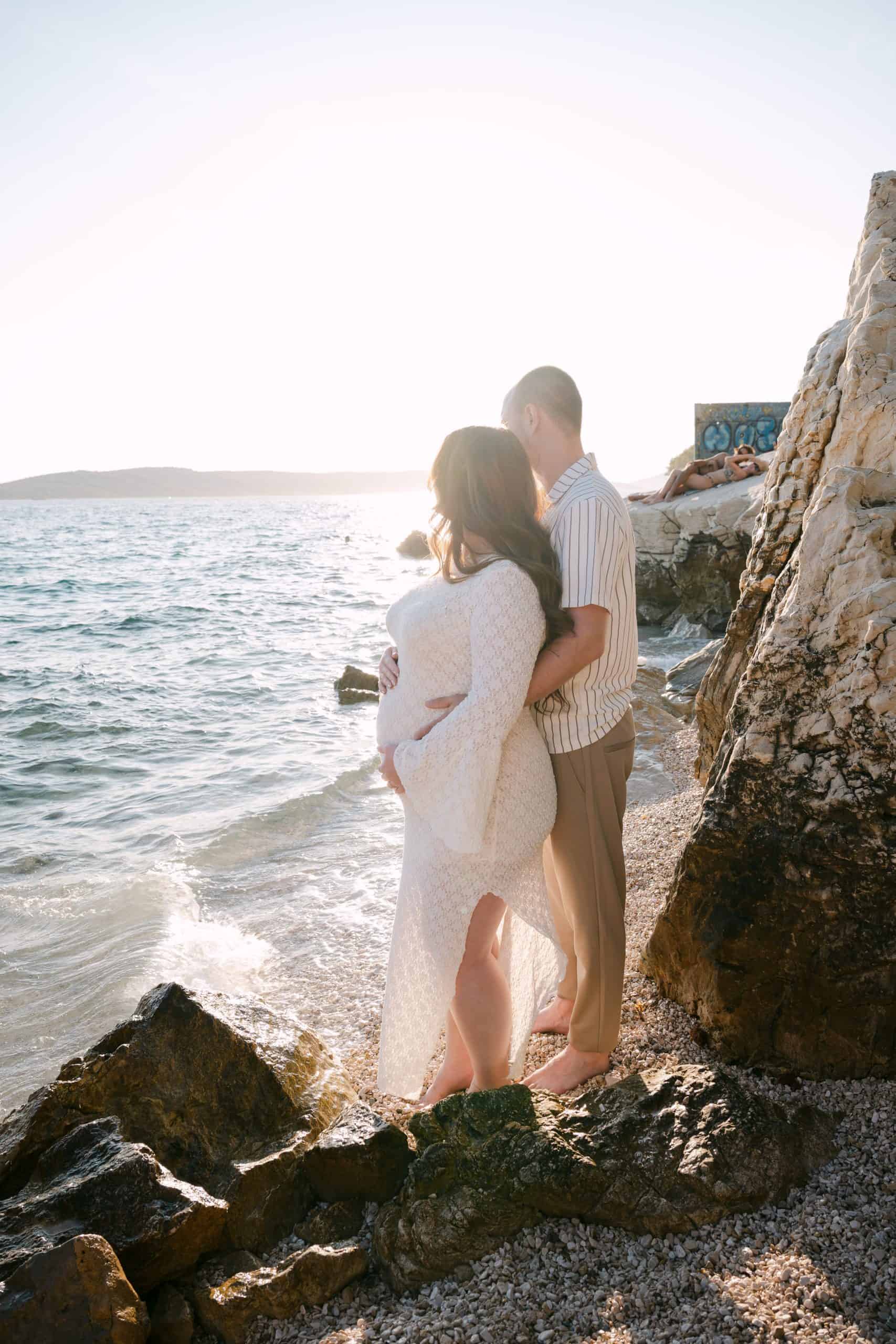 Zalazak sunca u uvali Obojena: romantični portret trudnog para uz šum mora i stijene.