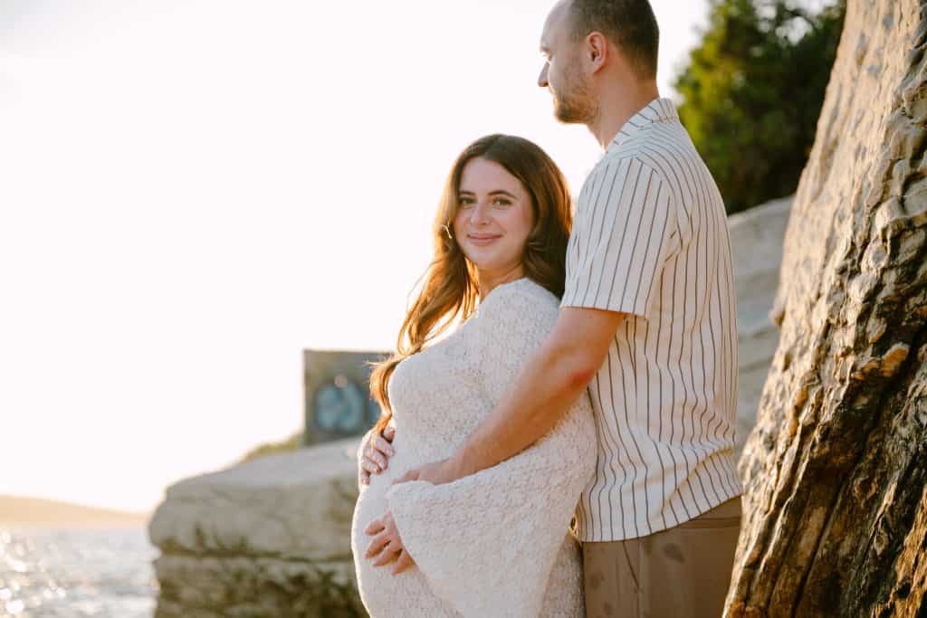 Trudnica i partner se grli na plaži tijekom zalaska sunca, bilježeći ljubav i povezanost.