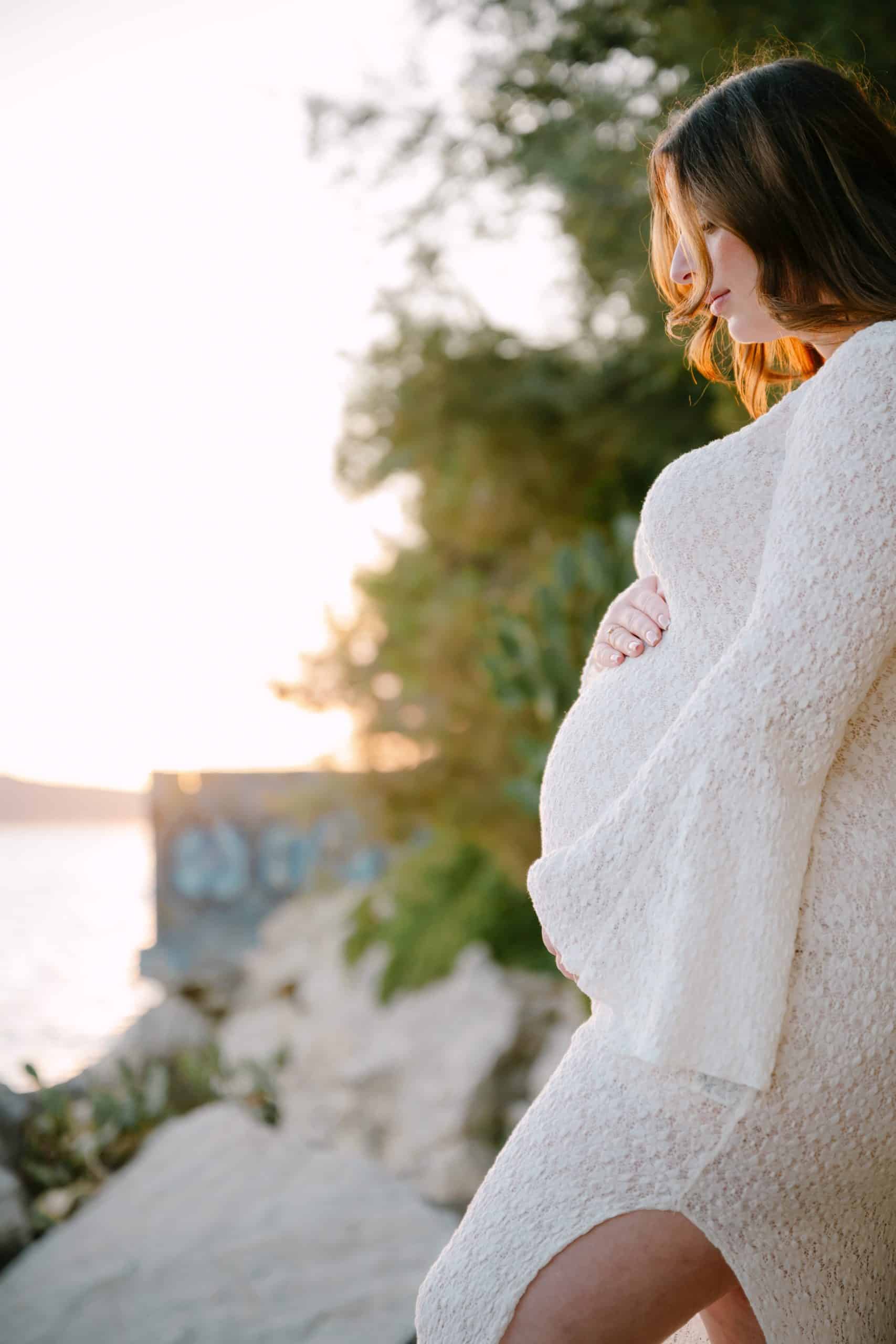 Trudna majka u bijeloj haljini na obali mora tijekom zalaska sunca.