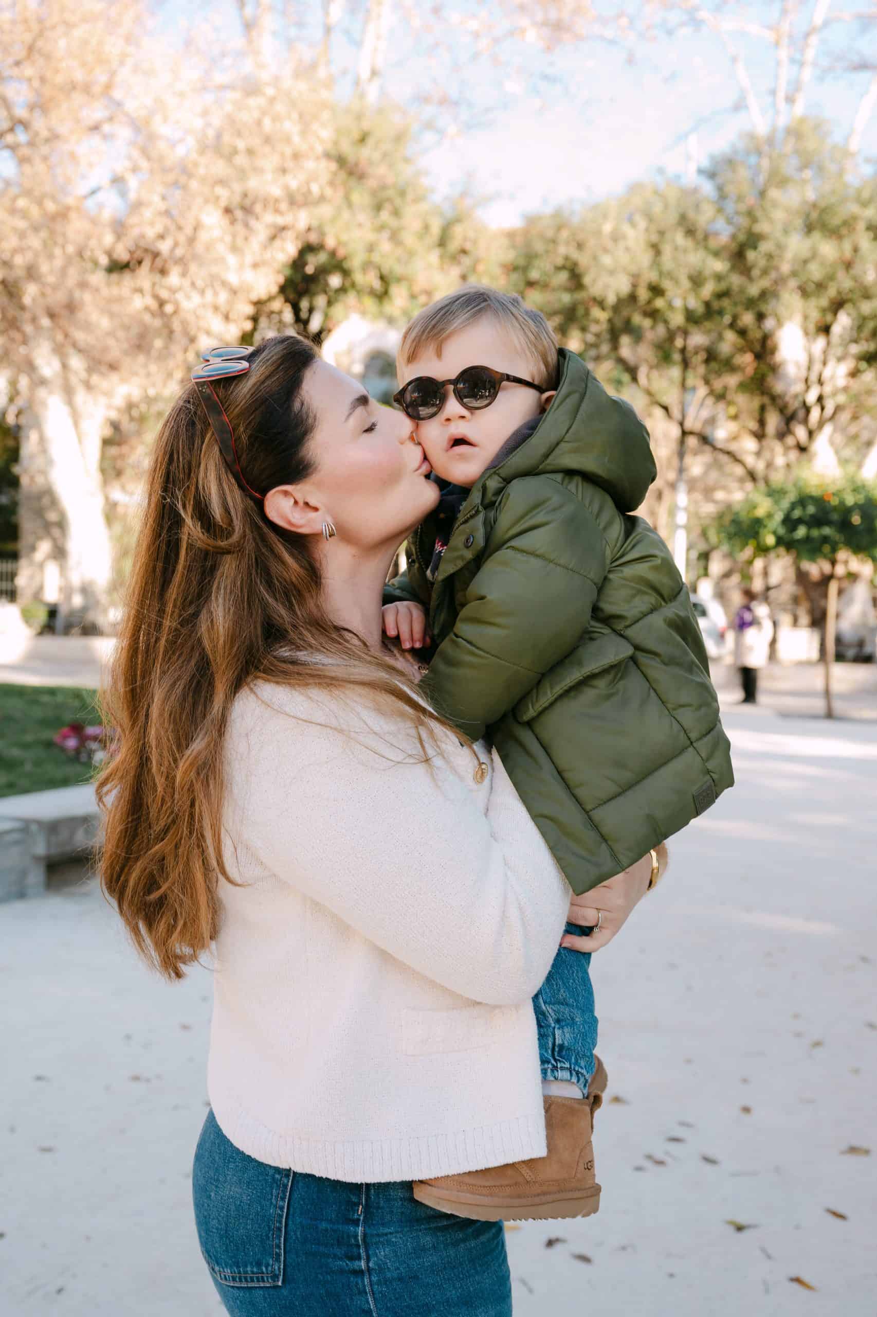 Majka drži sina, oboje toplo odjeveni, s drvećem i sunčevom svjetlošću u pozadini.