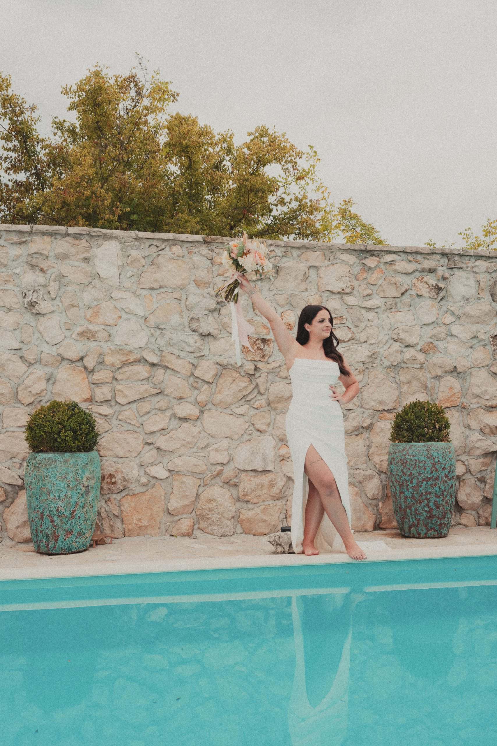 Portret mladenke u modernoj vjenčanici s buketom u ruci pored tirkiznog bazena. Elegantna vjenčana fotografija u luksuznom mediteranskom stilu.