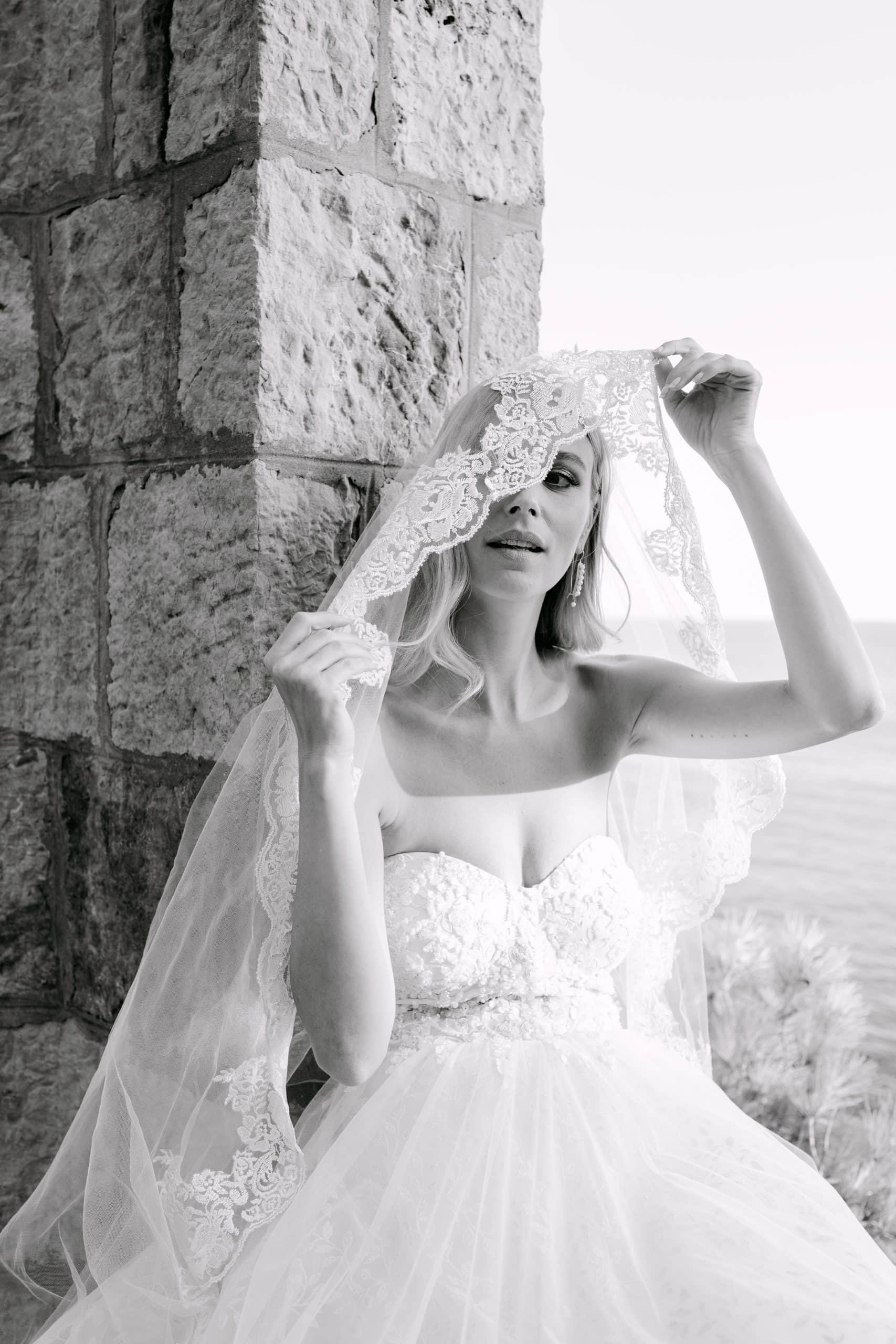 Filmski crno-bijeli portret mladenke koja rukama elegantno drži rubove svog čipkastog vela preko glave, pozirajući uz stari kameni zid u Dubrovniku.