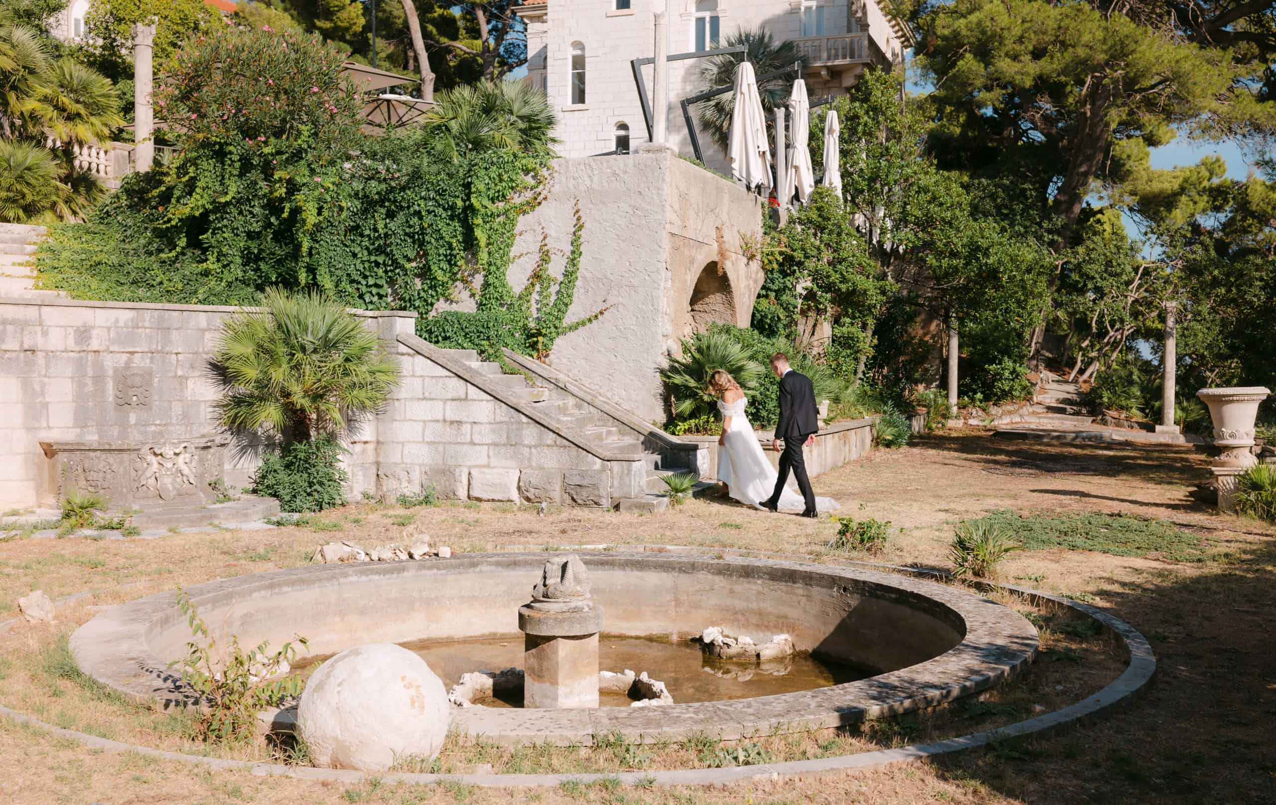 Mladoženja vodi mladenku za ruku u šetnji pored kružne kamene fontane u mediteranskom vrtu Vile Čingrija u Dubrovniku.