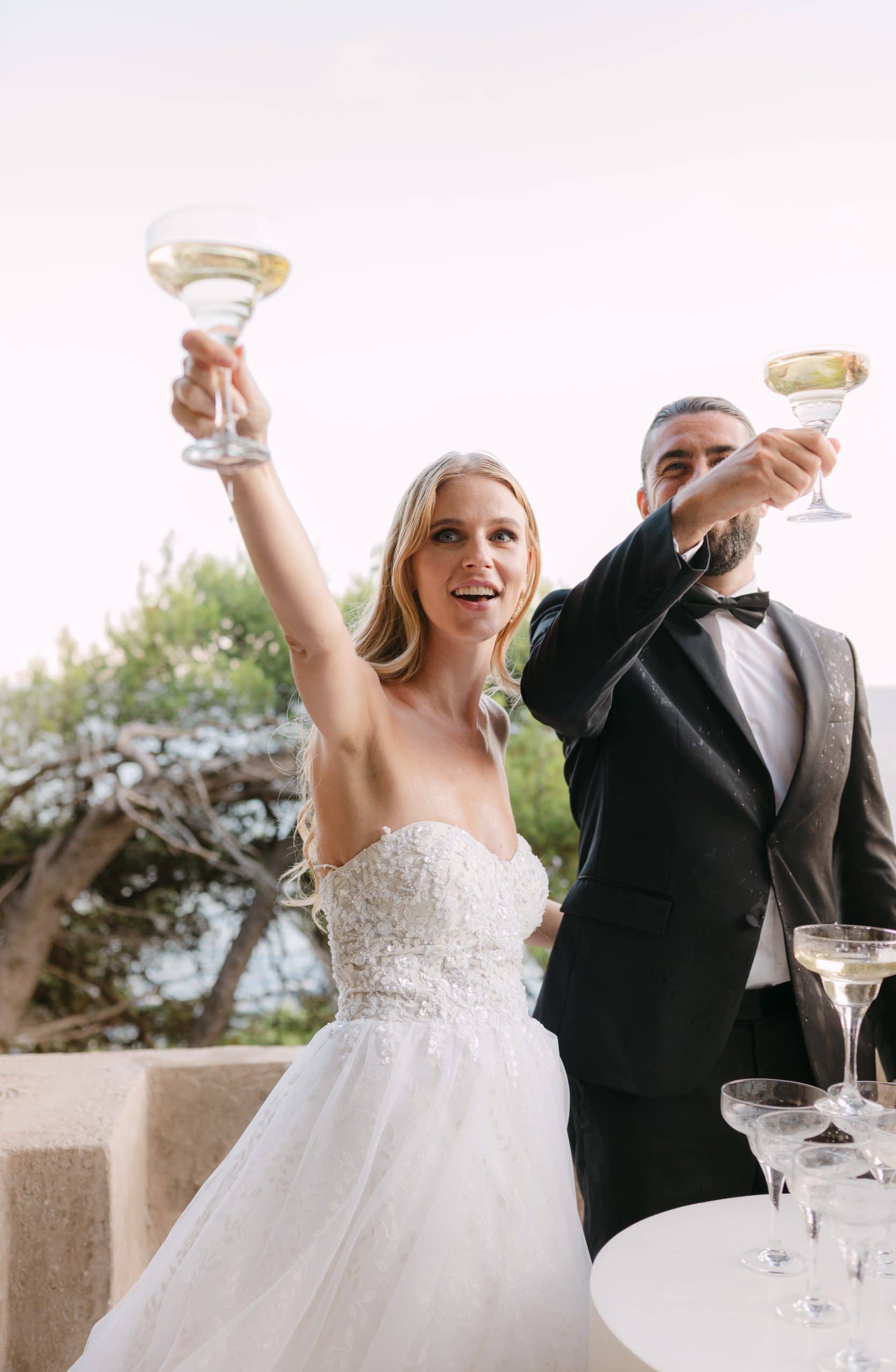 Mladenci veselo podižu čaše sa šampanjcem visoko u zrak slaveći svoje vjenčanje. Zabavna i spontana dokumentarna fotografija sa sessiona u Dubrovniku.